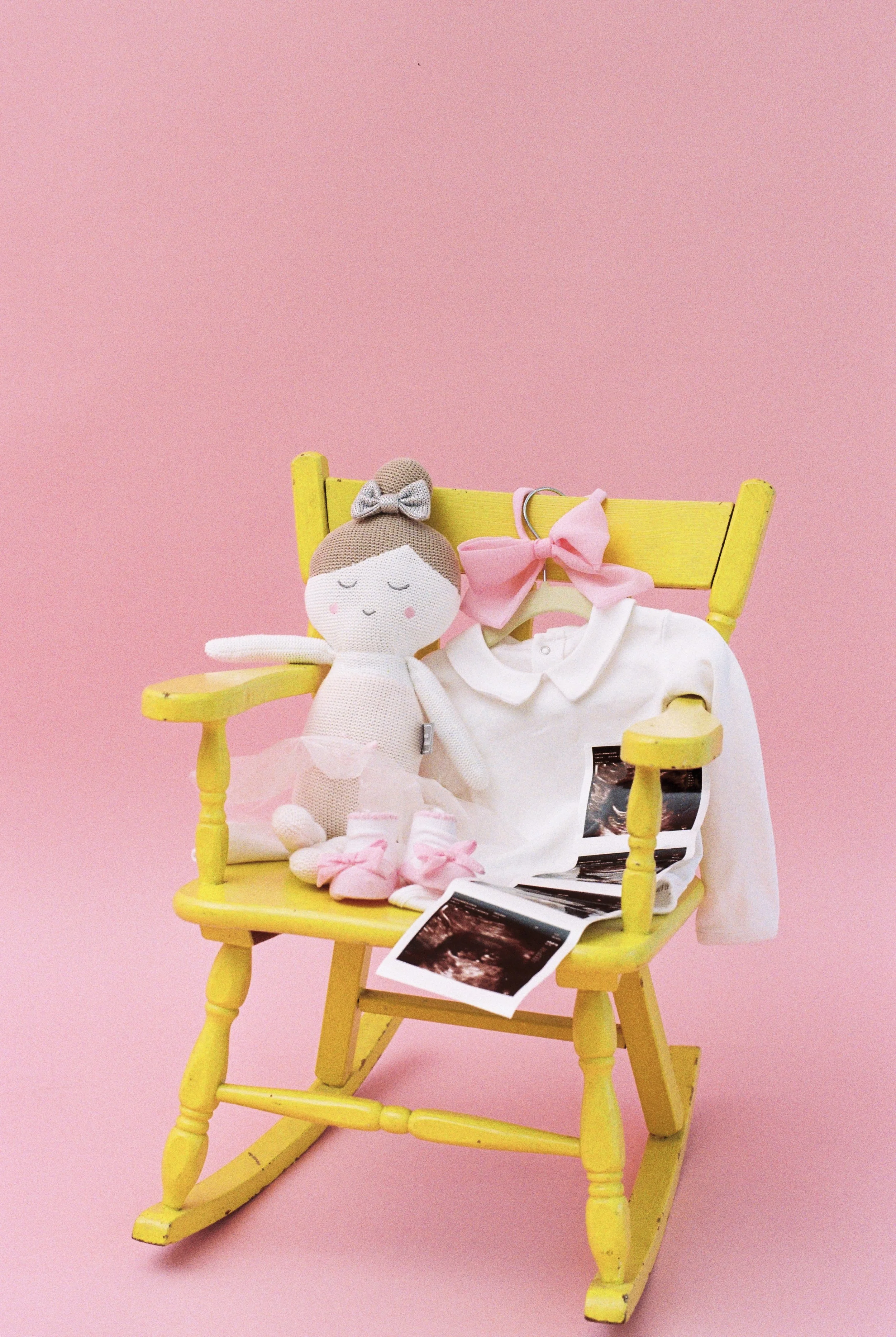 Bright yellow rocking chair against pink background with stuffed doll, baby clothes, ultrasound photos, and pink bows on it.
