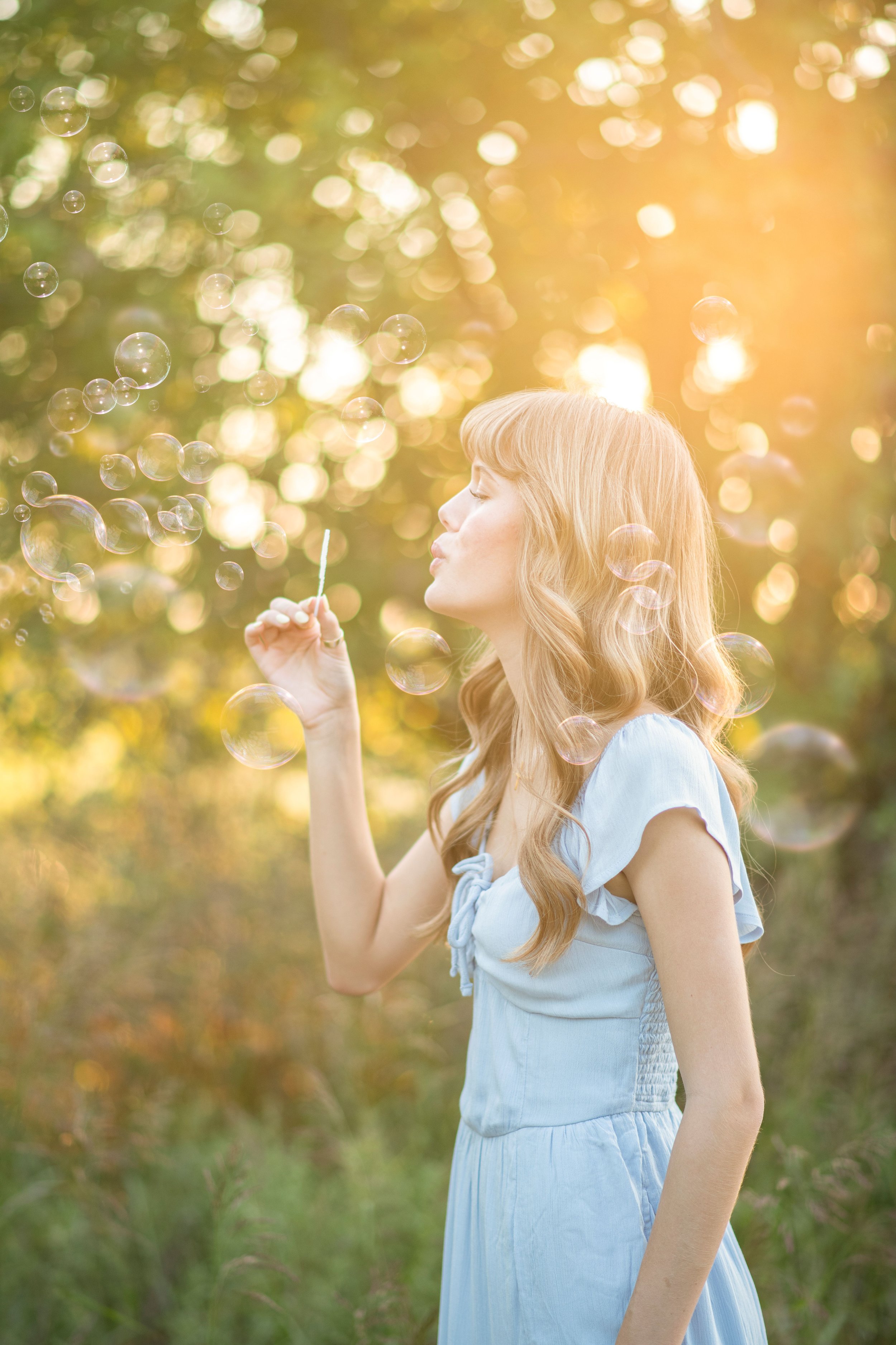 A young woman with long, wavy red hair wearing a light blue dress, blowing bubbles outdoors with sunlight filtering through trees in the background.
