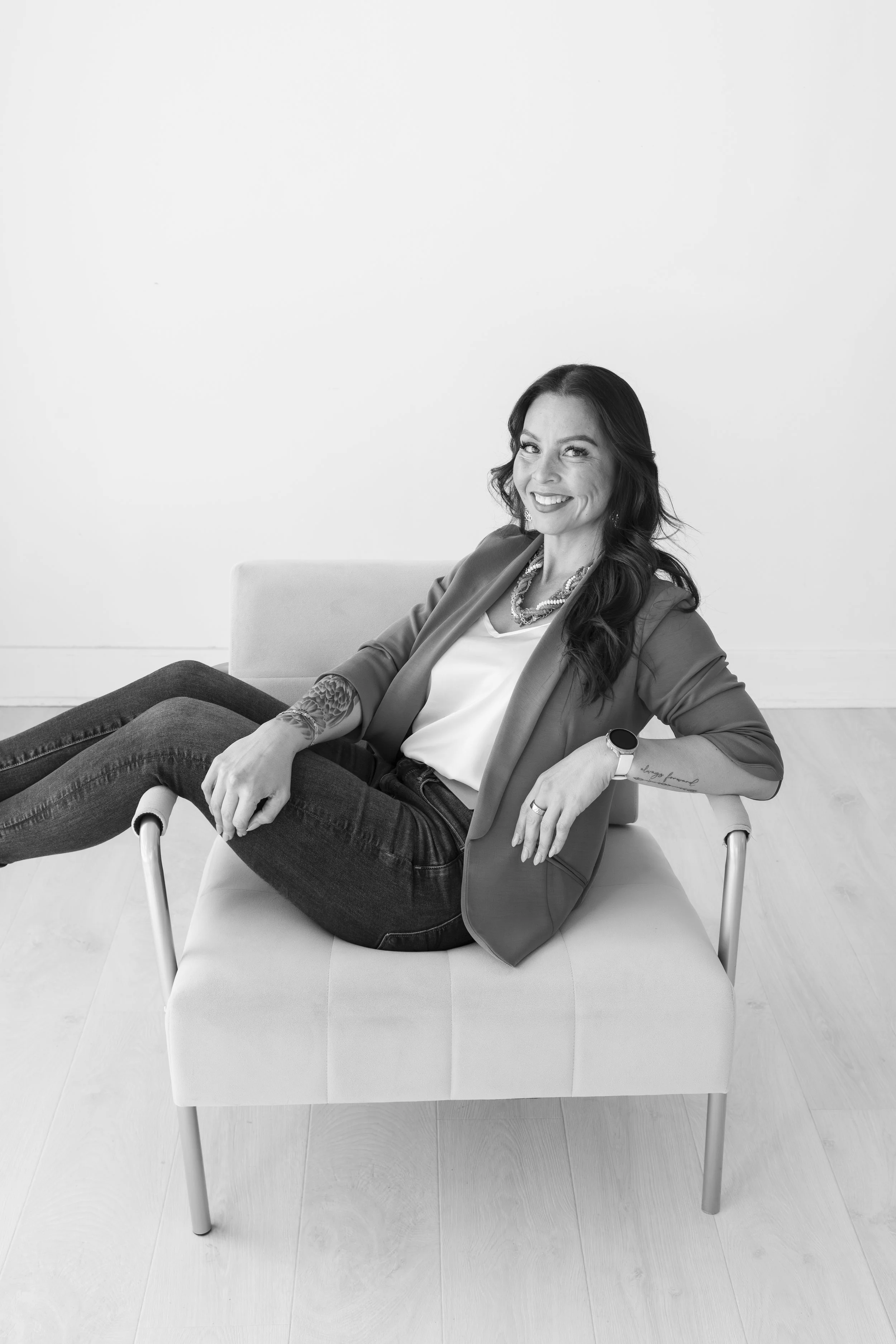 Black and white photo of a woman sitting on a chair with her legs crossed at the knee, smiling, wearing a blazer, white blouse, and dark jeans, with a tattoo on her arm, in a room with plain white wall and wooden floor.