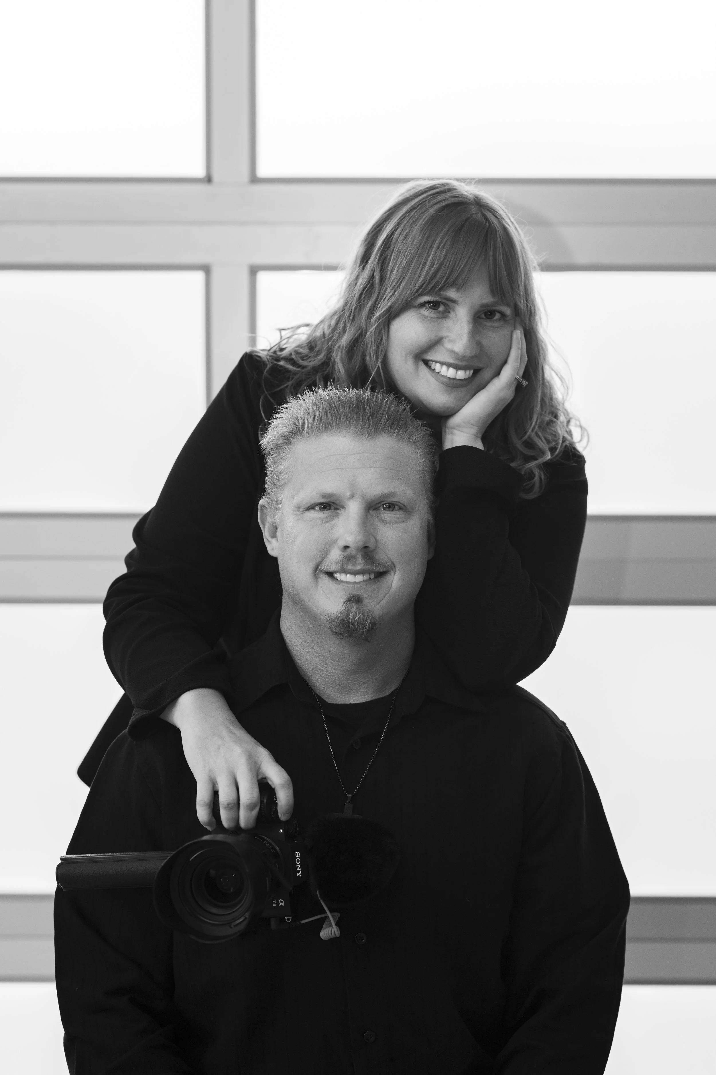 Black and white photo of a smiling woman with wavy hair leaning on a woman with short hair and a goatee, both indoors in front of a window, with a camera visible in the foreground.