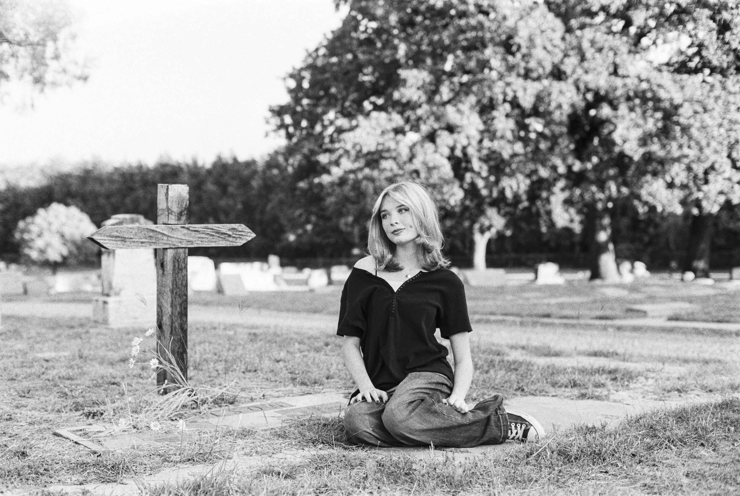A black-and-white photo of a woman sitting cross-legged on the grass in a park, near a weathered wooden signpost, with large trees and gravestones in the background.