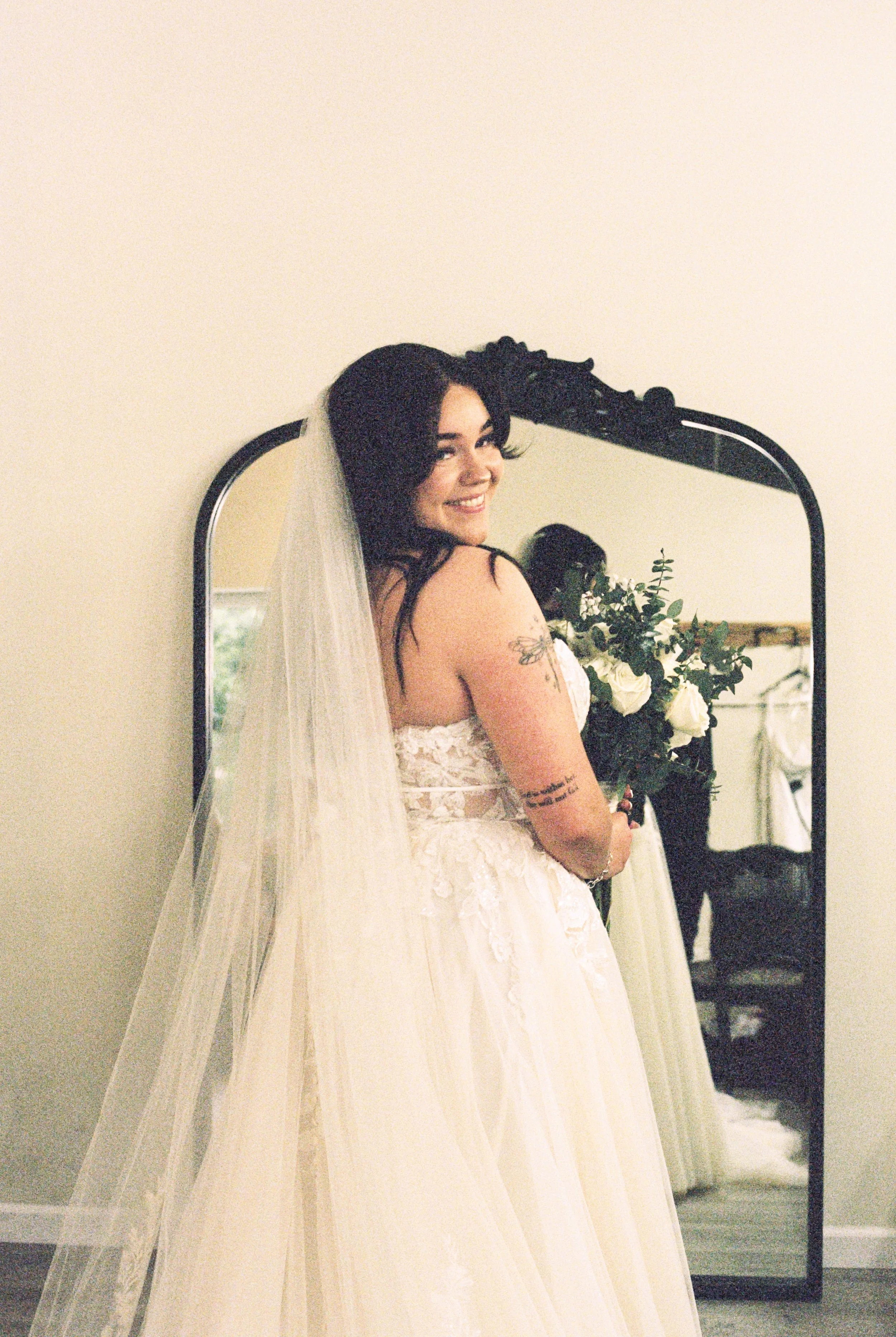 A bride with dark hair smiling, holding a bouquet of white roses and greenery, wearing a white lace wedding dress and veil, standing in front of a mirror in a room with a beige wall.
