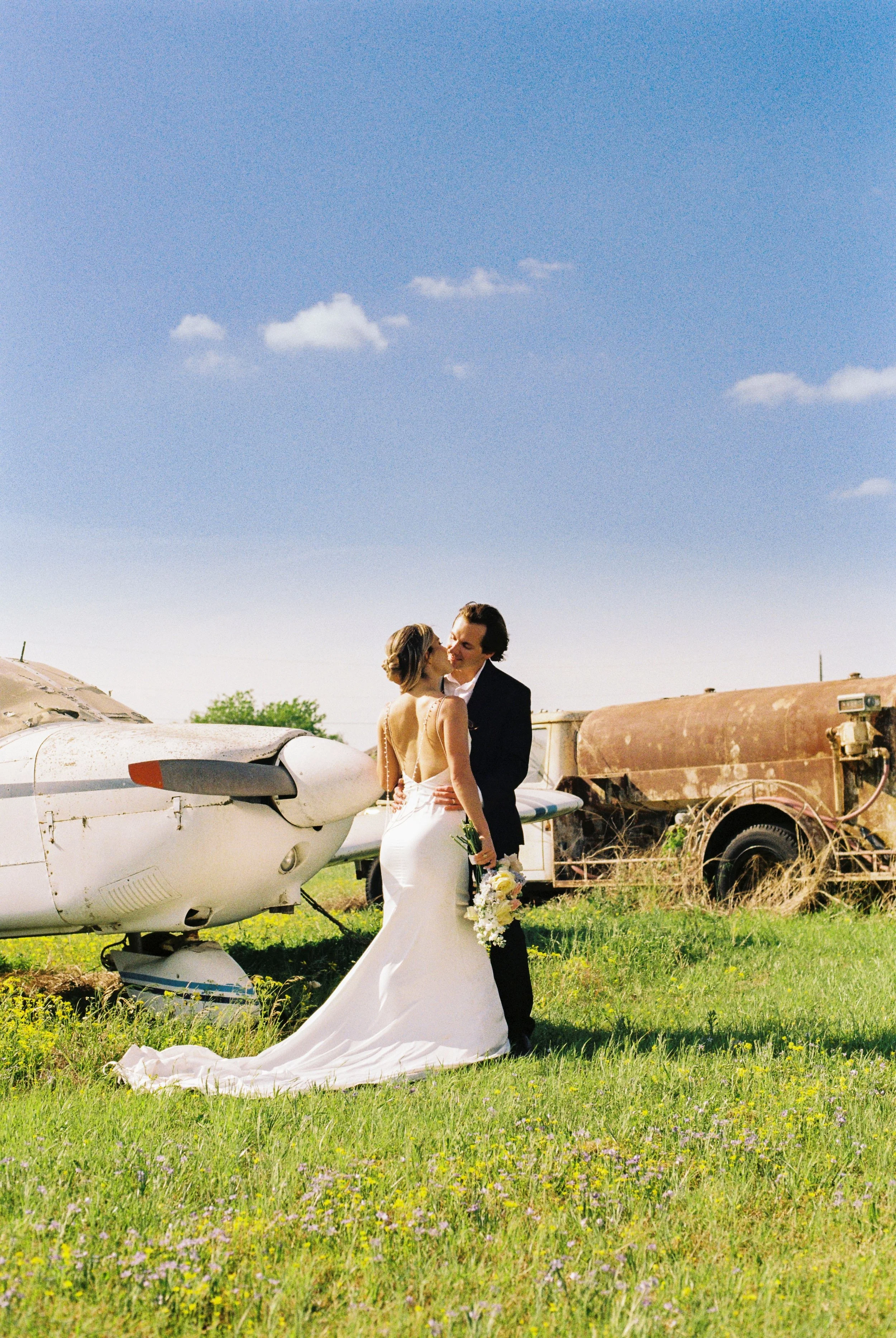 A bride and groom embrace outdoors beside vintage airplanes and rusted farm equipment under a bright blue sky.