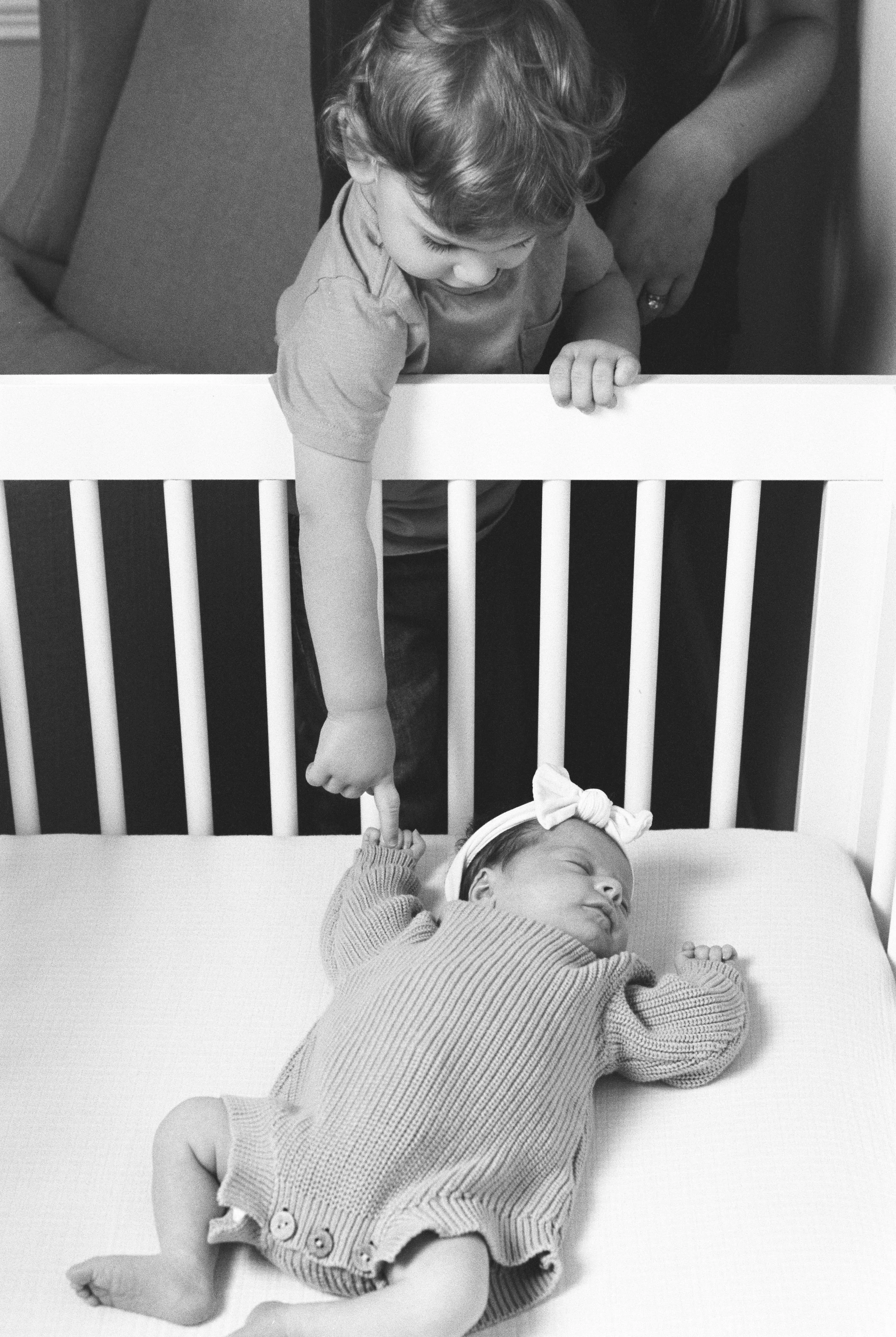A young boy leaning over a crib, pointing at a sleeping baby girl who is lying on her side with a headband and a ribbed bodysuit