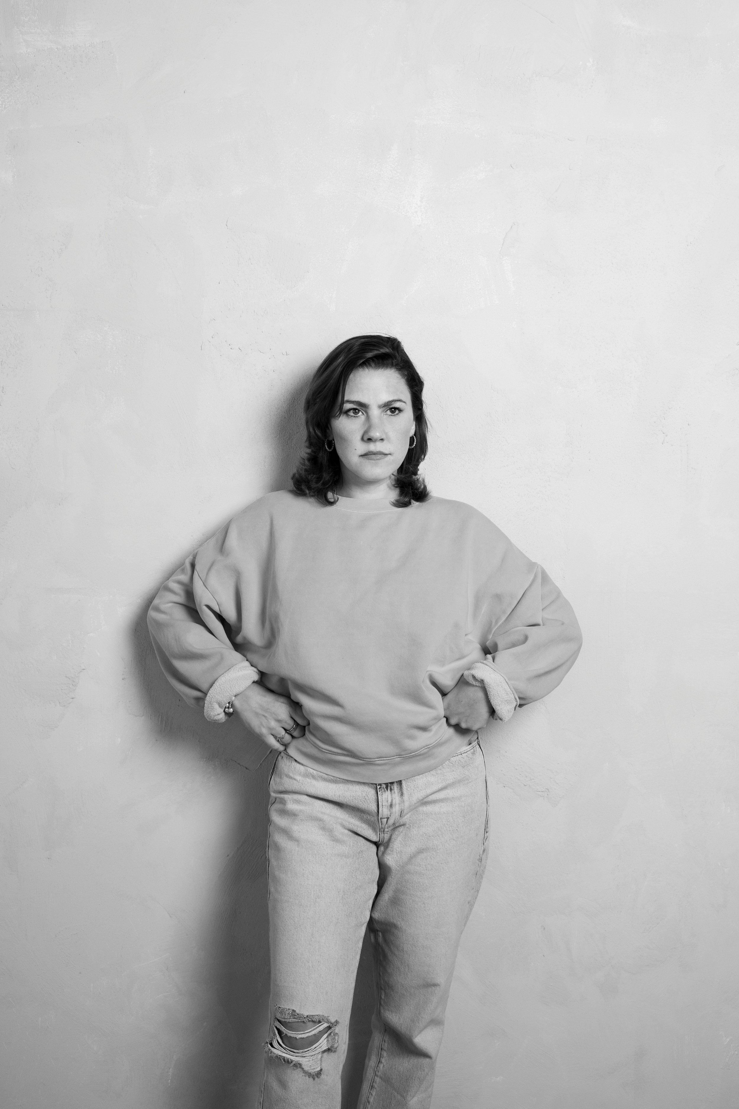 Black and white photo of a woman standing against a plain wall, with her hands on her hips, wearing a sweatshirt and ripped jeans.