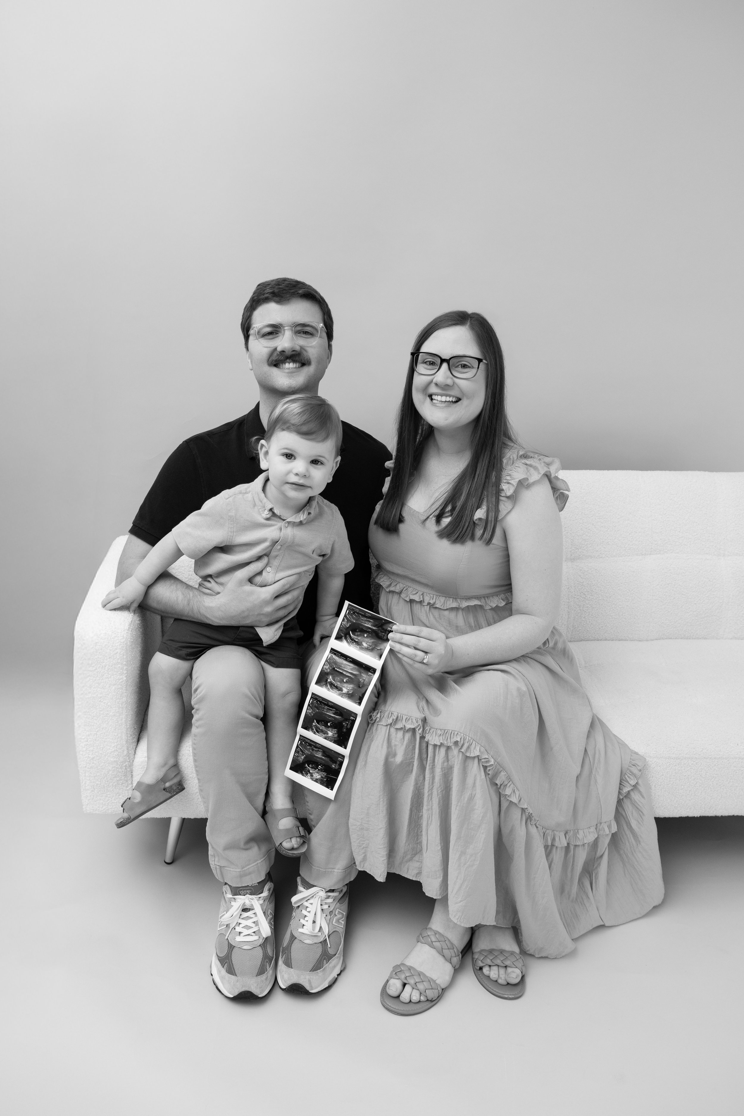 A black and white photo of a family of three sitting on a couch. The man has a mustache and glasses, and the woman has long hair and glasses. The young boy is sitting on the man's lap, holding ultrasound images. All are smiling.