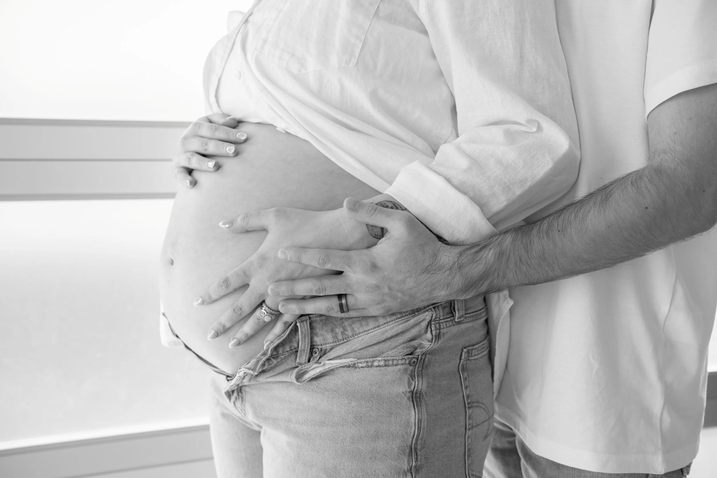 A pregnant woman with her partner touching her belly. The woman is wearing jeans and a shirt, the partner is wearing a ring and a watch.