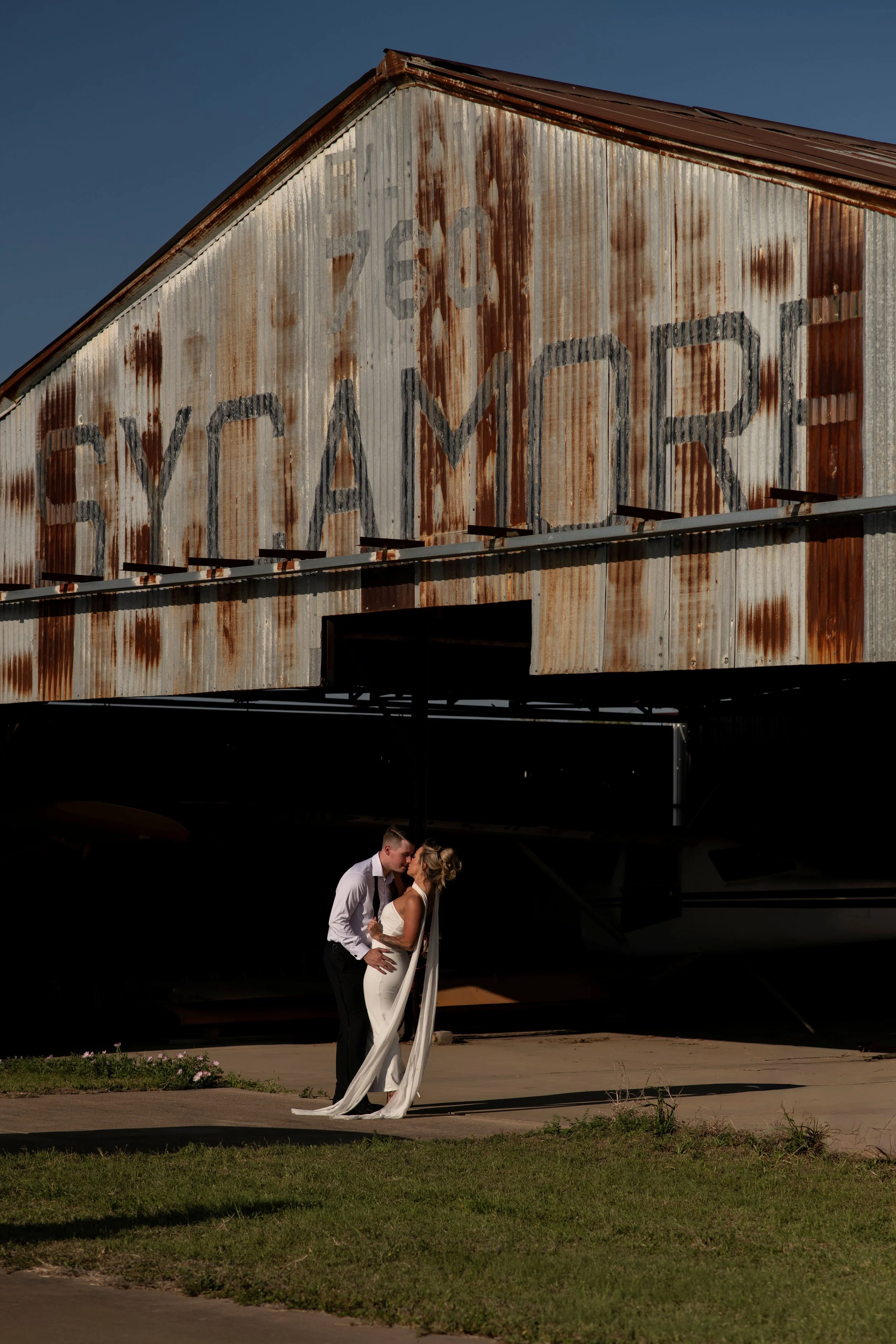 A couple dressed in wedding attire standing close together outdoors, with a rustic, weathered metal barn in the background featuring large faded paint labels.