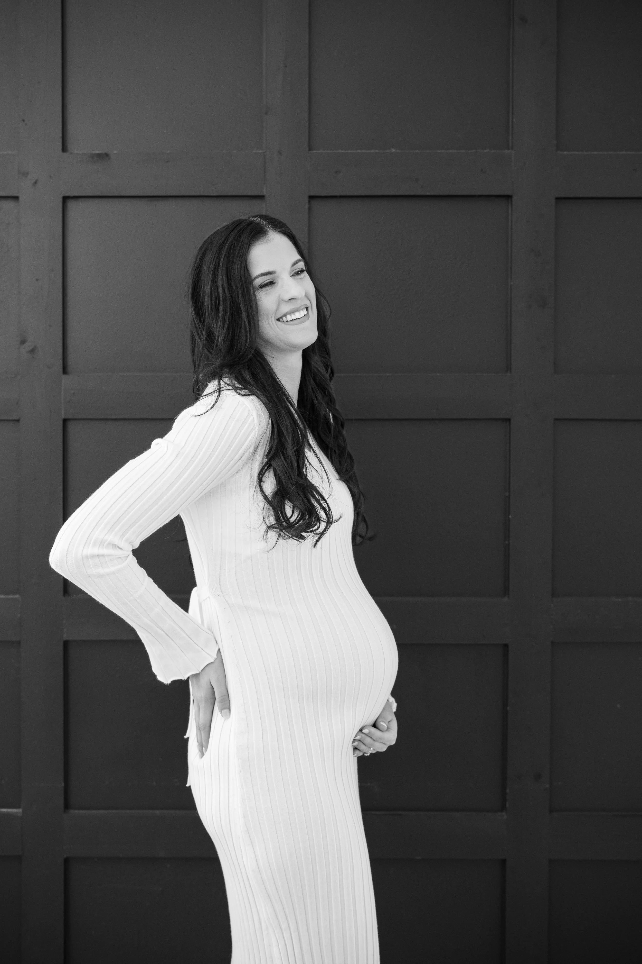 Black and white photo of a smiling pregnant woman in a long, ribbed dress with her hands on her hips and her belly, standing in front of a wooden panel wall.