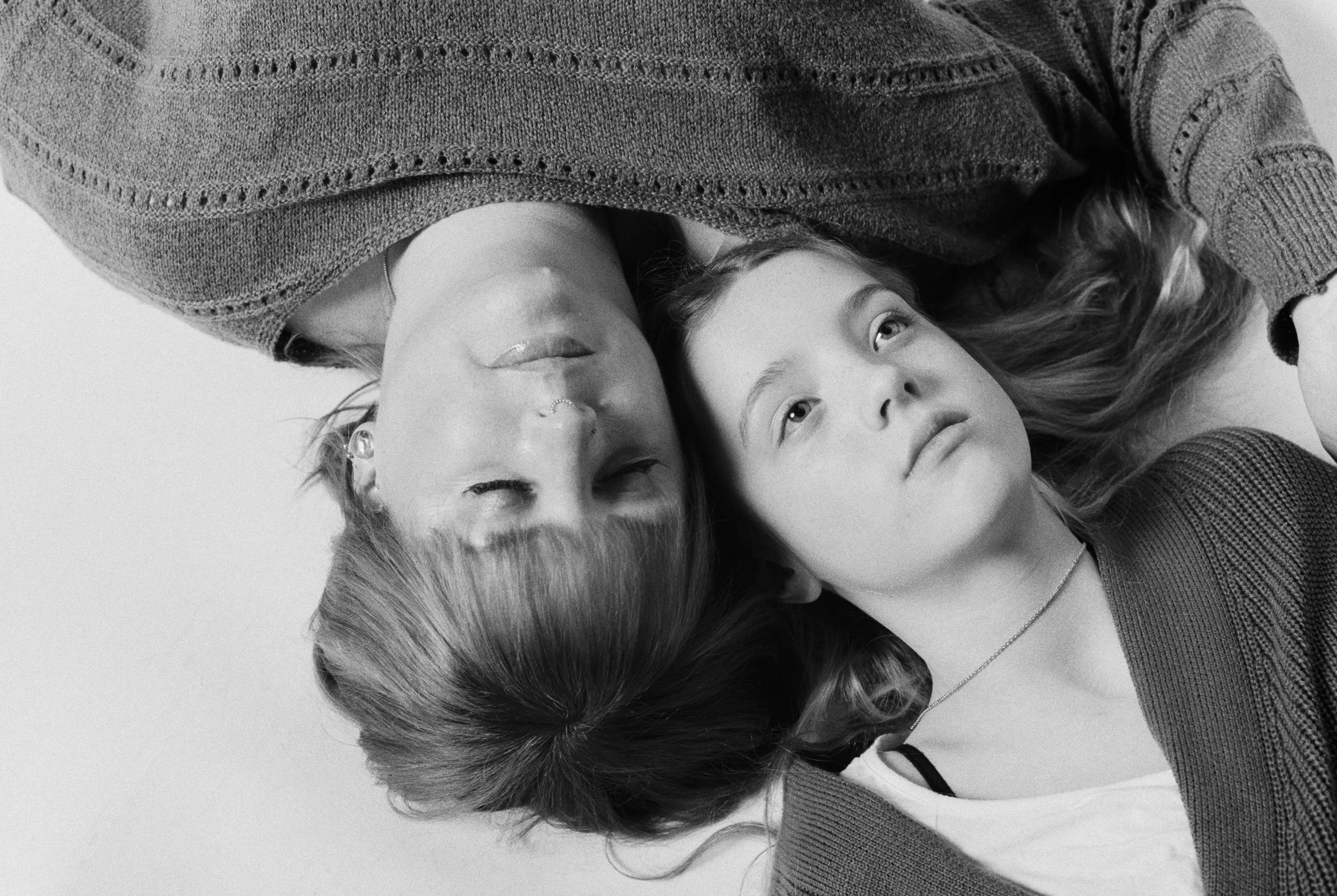 Black and white photo of two women lying down with their heads close together, looking upwards. One woman has her hair covering part of her face and is wearing a textured sweater. The other woman has light hair, a necklace, and is also wearing a swea