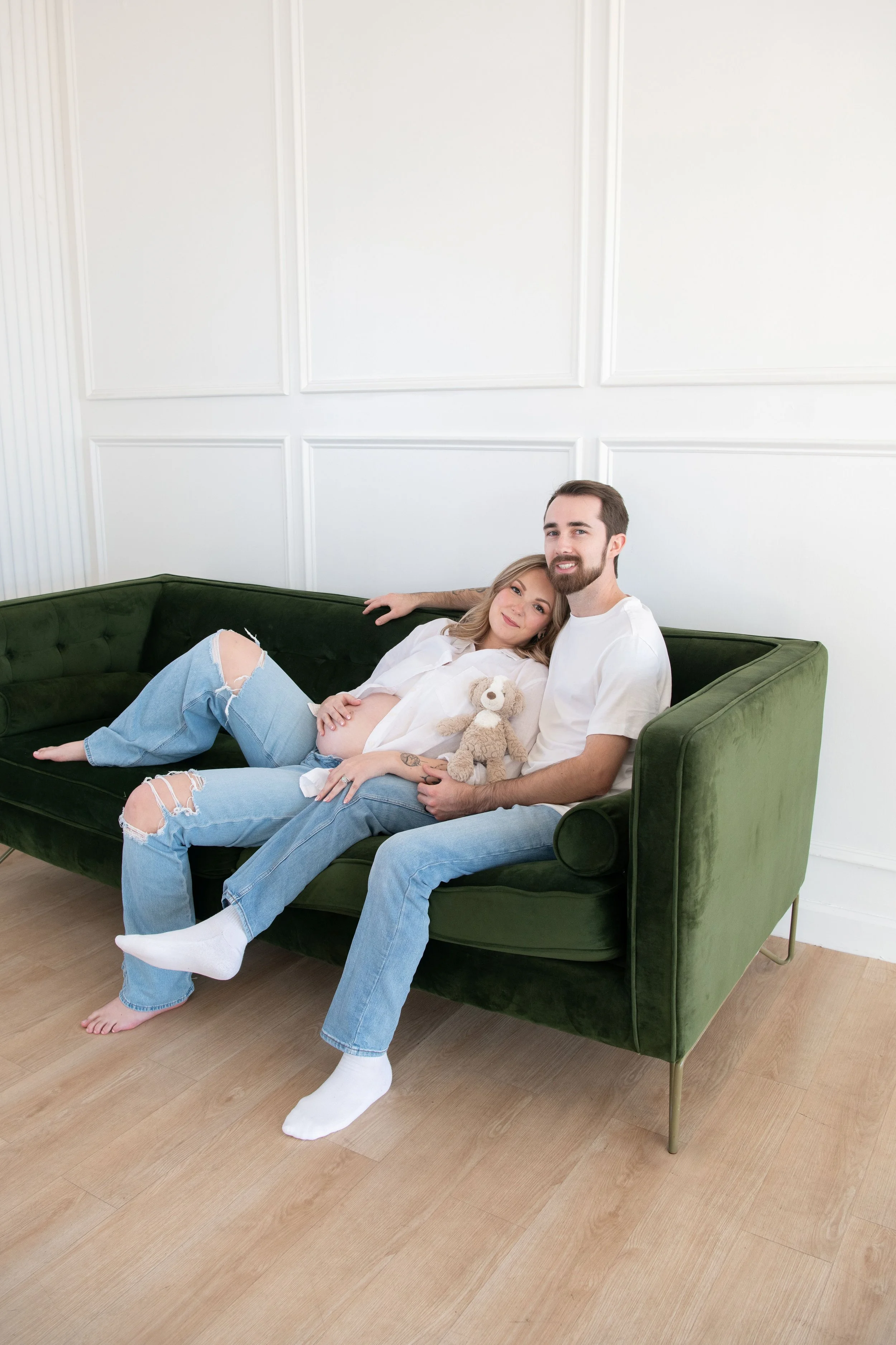 A pregnant woman and a man sit on a green velvet couch in a bright, minimal living room. The woman rests her head on the man's shoulder, holding a teddy bear, as they smile at the camera.
