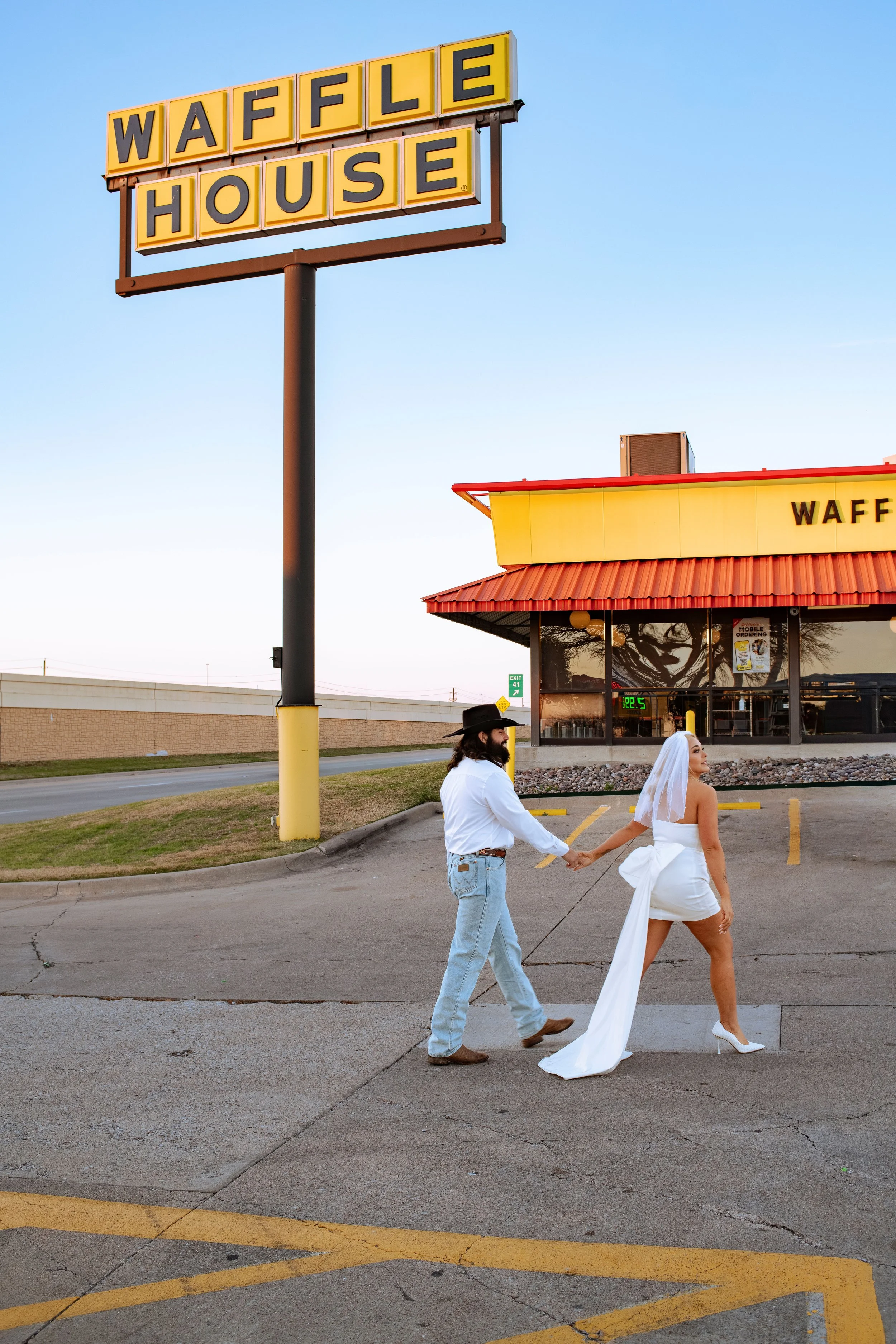A man and a woman in wedding attire holding hands and walking in a parking lot outside a Waffle House restaurant. The man is wearing a white shirt and jeans, the woman is wearing a white wedding dress and high heels.