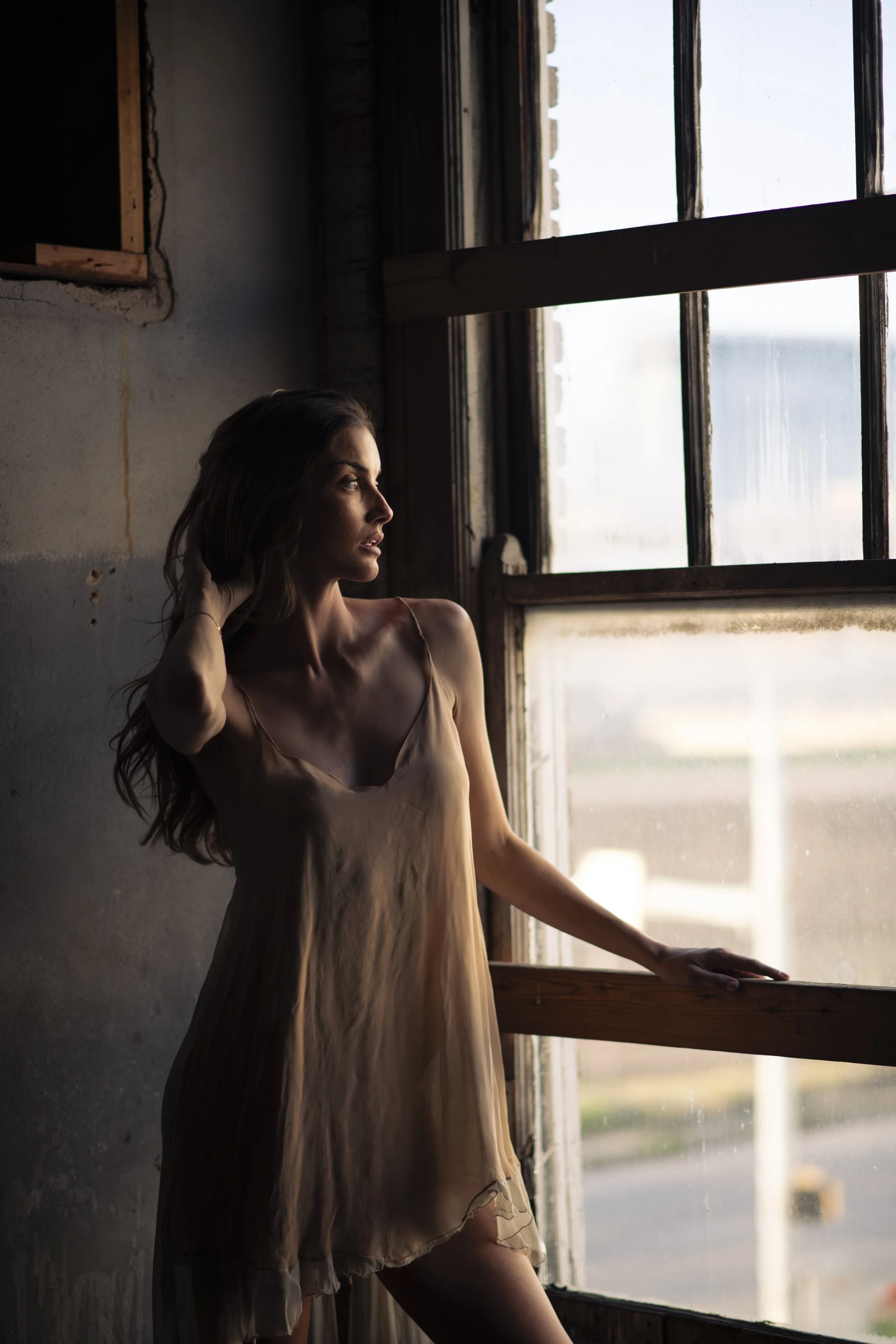 A woman with long brown hair wearing a beige dress standing by a window, looking outside, with sunlight illuminating her profile and casting shadows inside a rustic room.