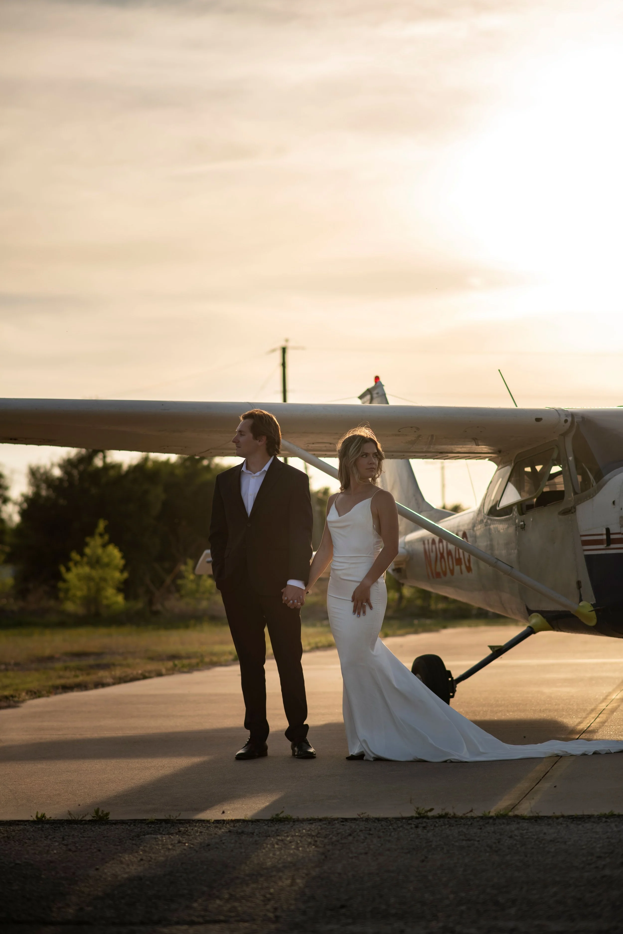 A man and woman dressed in formal wedding attire holding hands, standing beside a small airplane on a runway at sunset.
