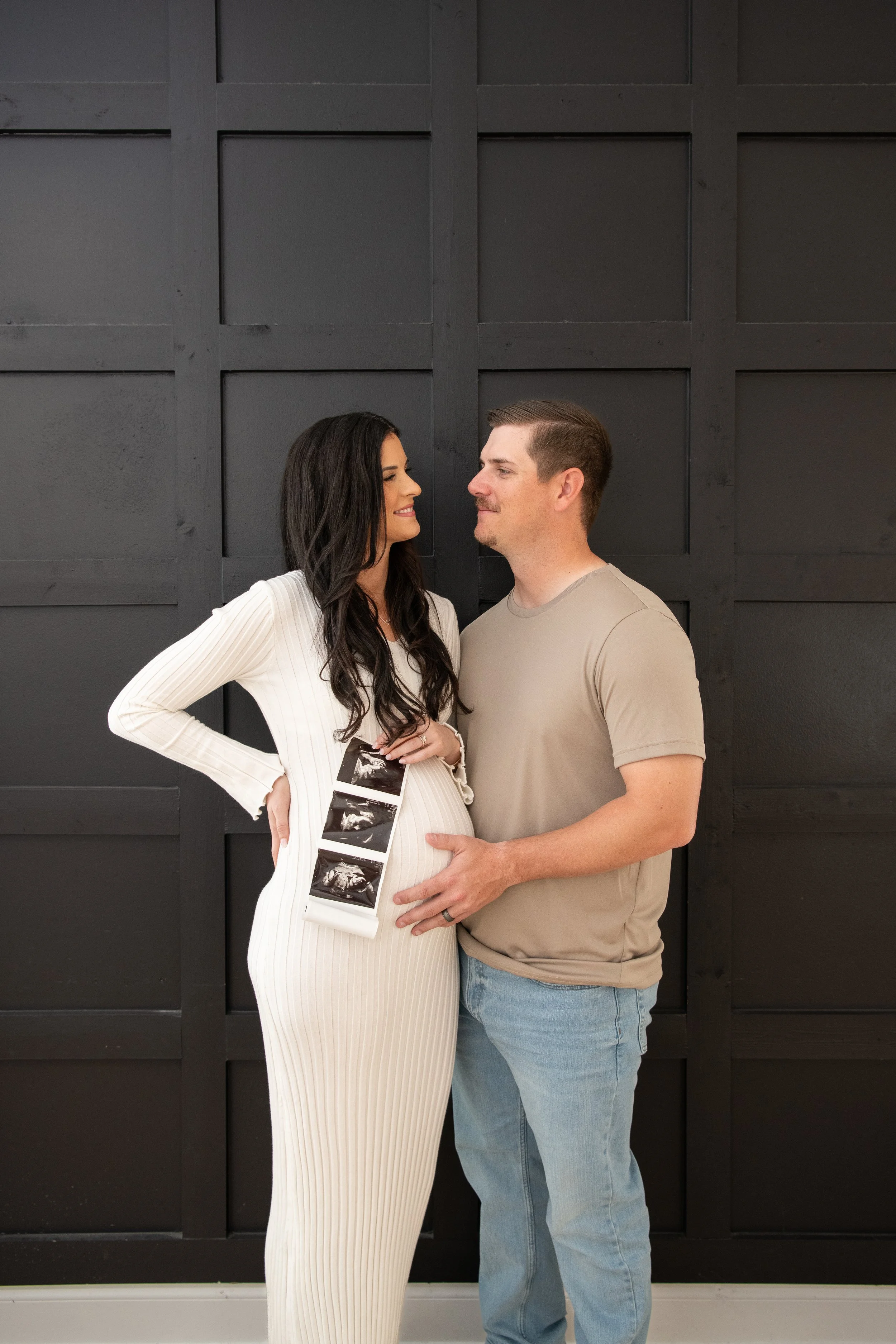 A couple standing in front of a black wall, with the woman displaying ultrasound images on her pregnant belly and the man touching it, both looking at each other lovingly.