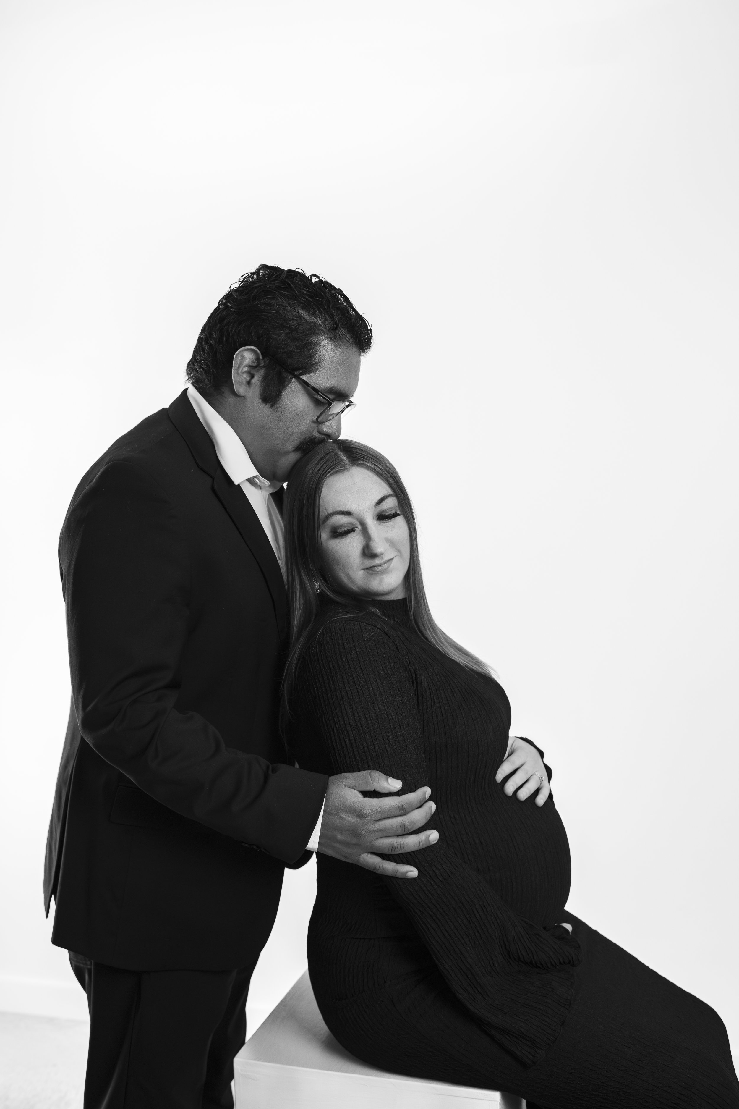 A black and white photo of a man in a suit with glasses kissing a pregnant woman on the head. The woman, wearing a long-sleeved black dress, is sitting on a white surface with a tender expression, holding her baby bump. The man is standing beside her