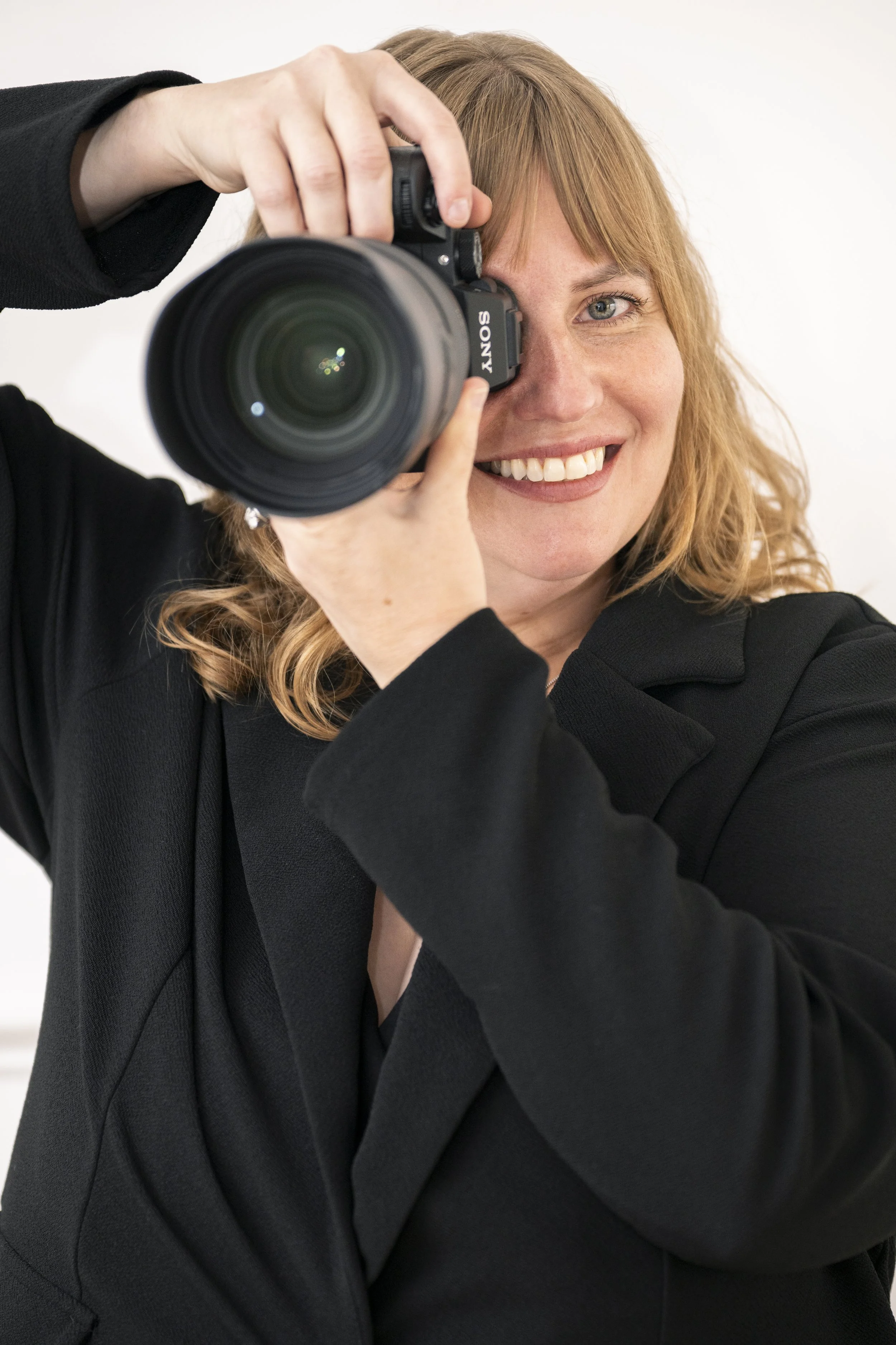 A woman with red hair in a black blazer smiling while taking a photograph with a Sony camera.
