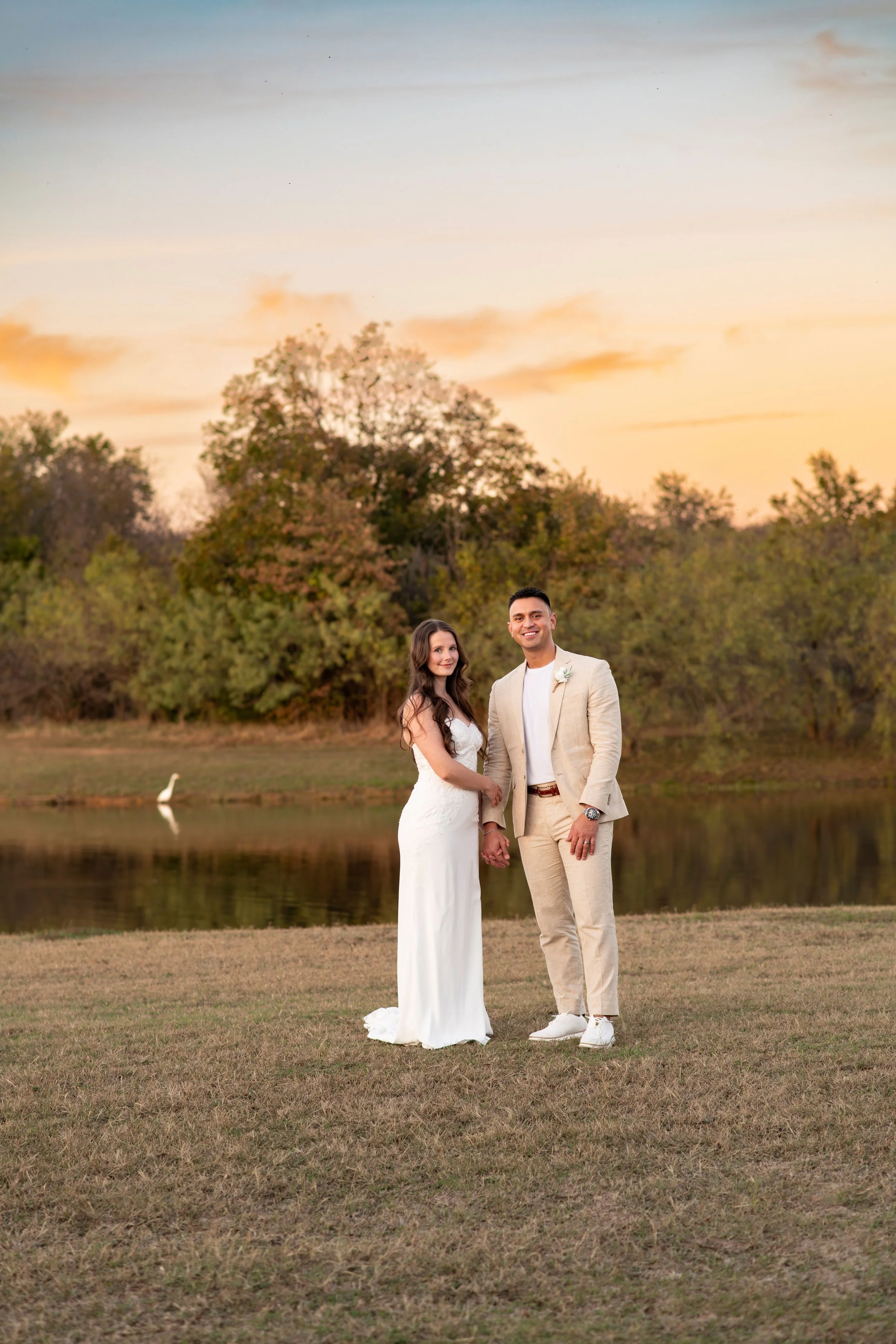 A couple dressed in wedding attire standing hand-in-hand outdoors by a lake, with a white swan swimming in the background and a sunset sky overhead.