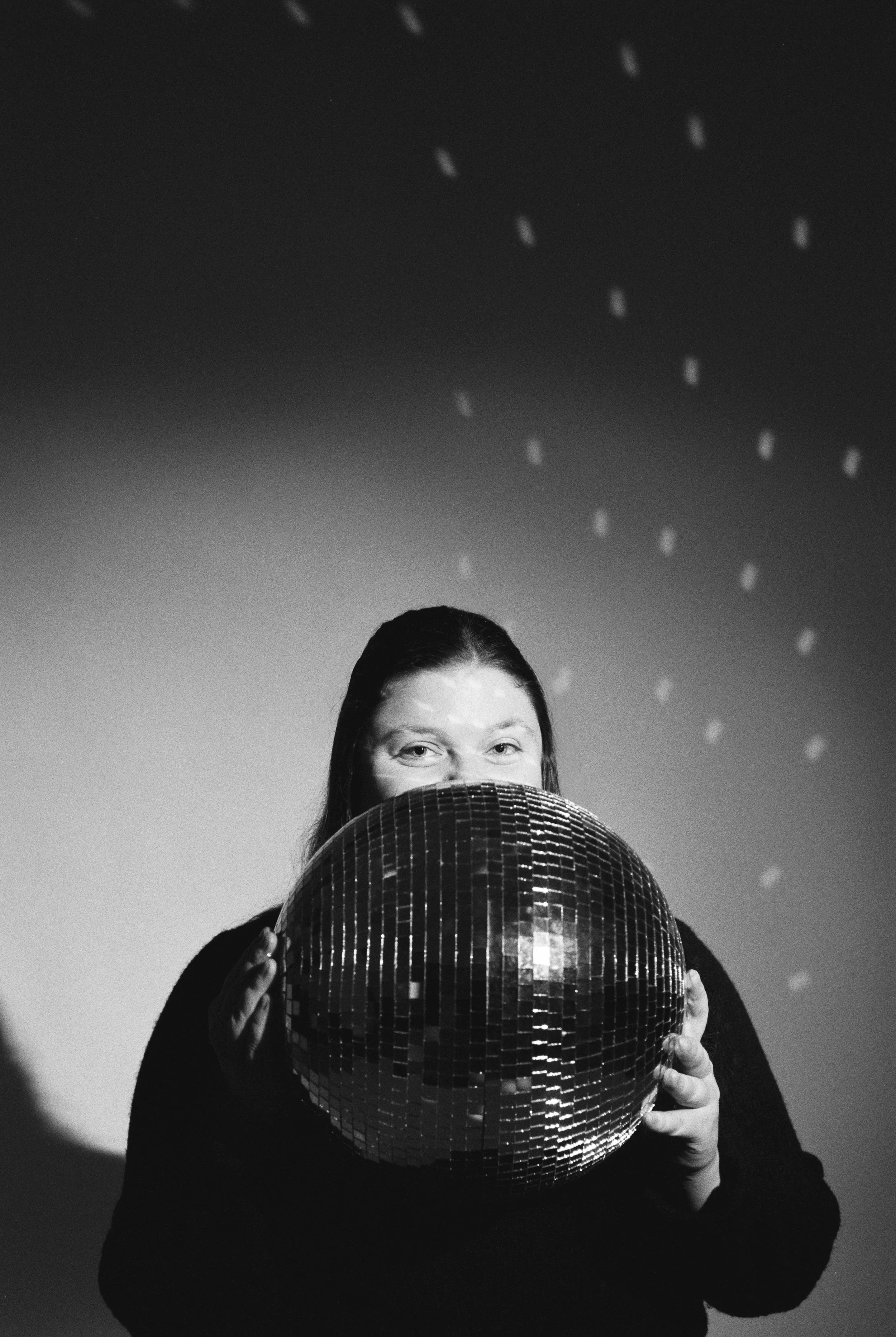 A person holding a reflective disco ball covering their mouth, with a gradient background and small lights projected onto the wall.