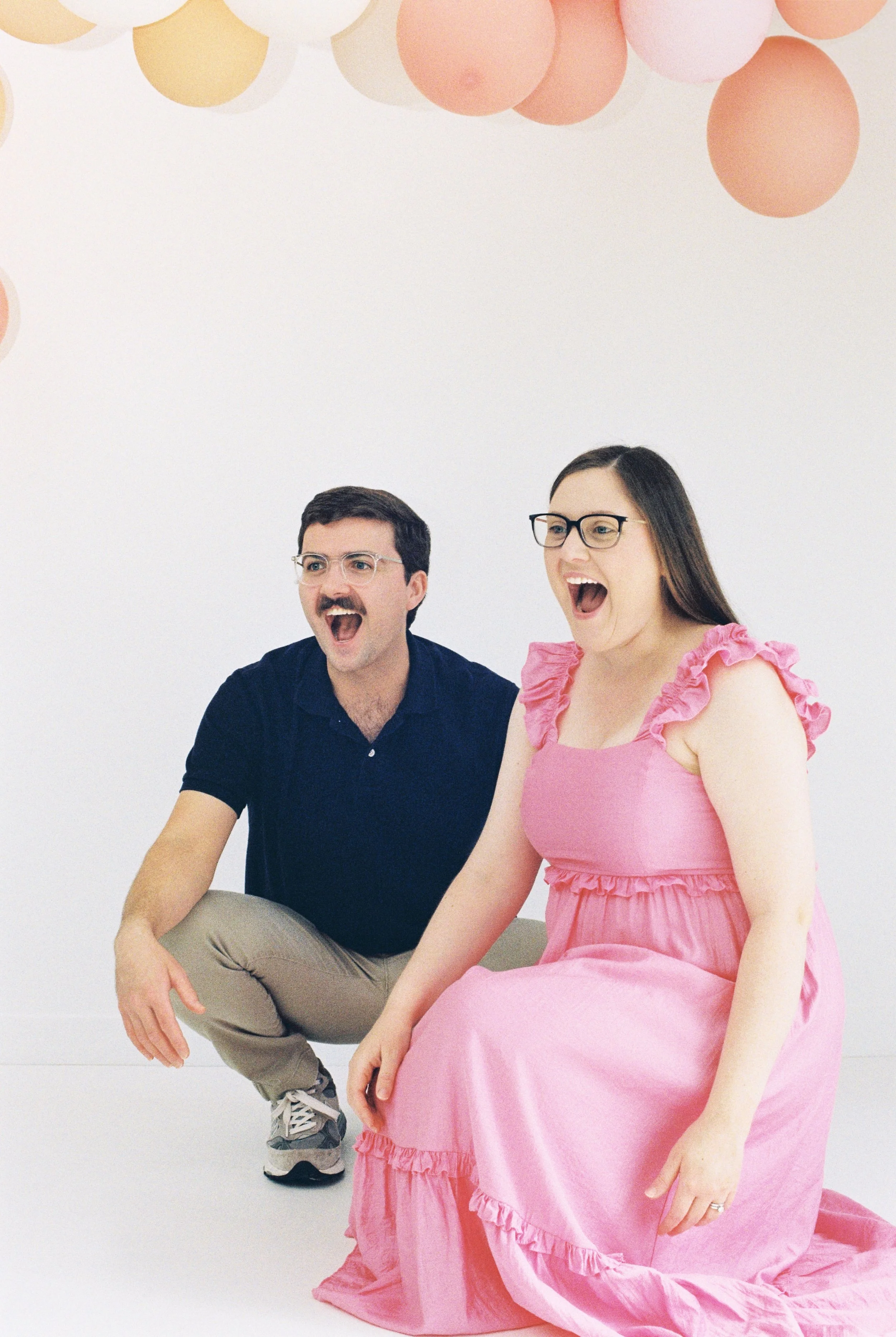 A man and a woman are seated against a white background decorated with pink and peach paper balloons, both appear to be surprised or excited.