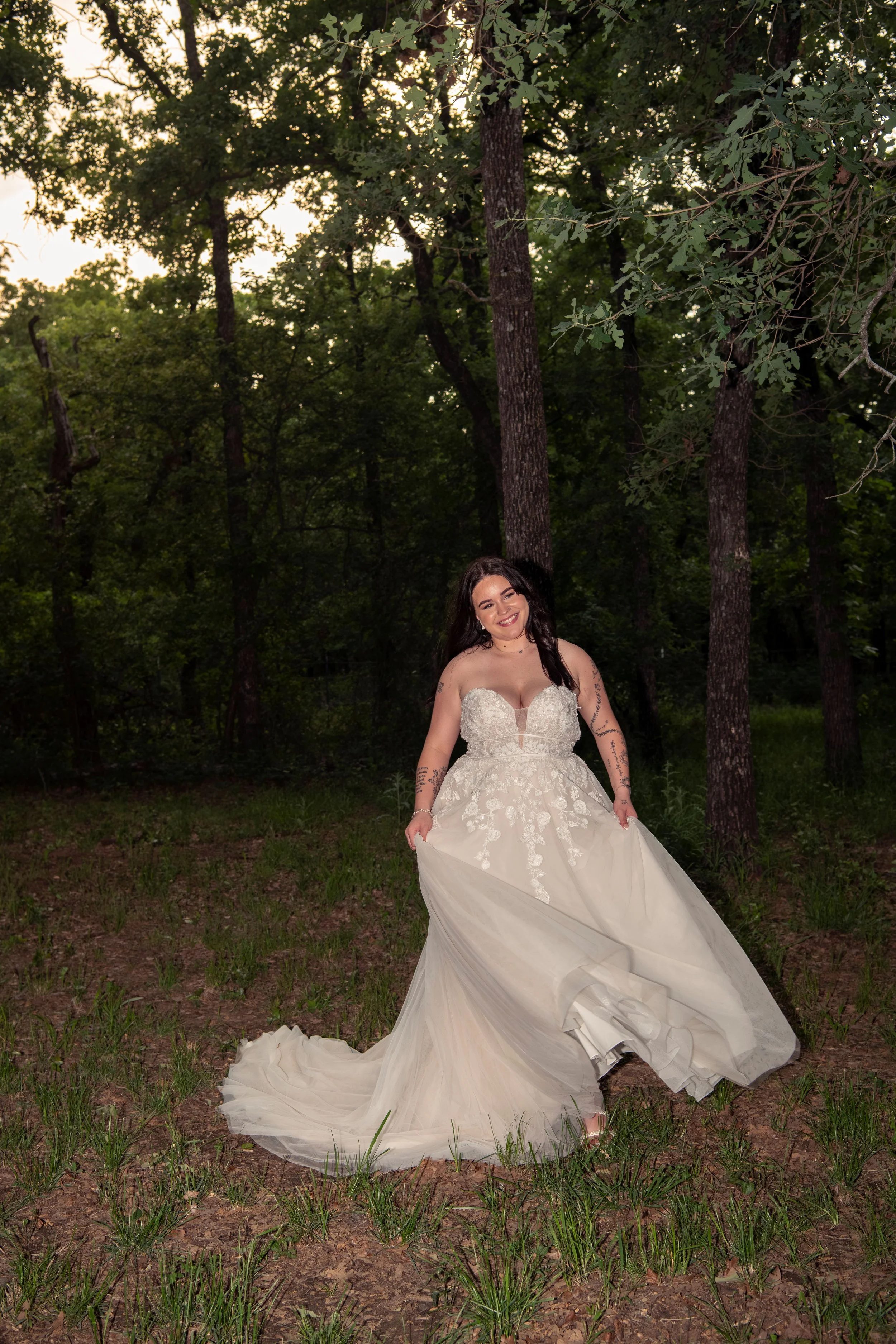 A woman in a white wedding gown standing outdoors in a wooded area, smiling, with trees and evening sky in the background.