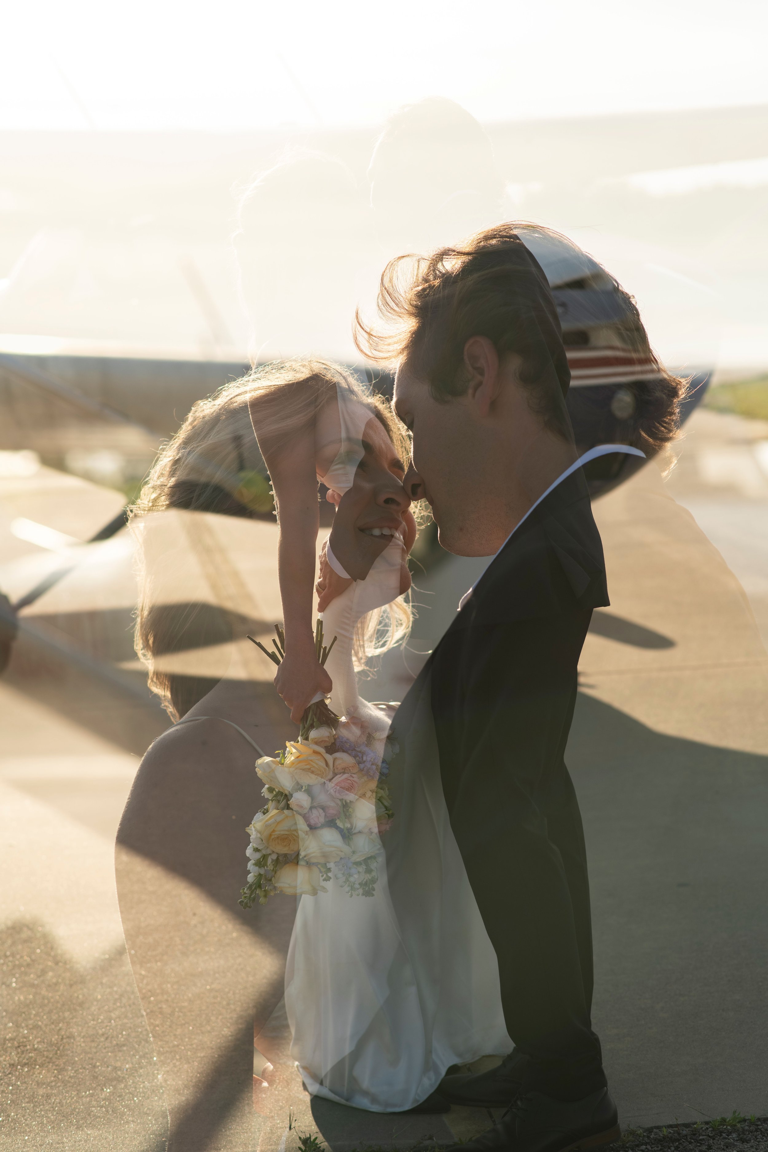 A couple in wedding attire sharing an intimate moment outdoors, with the groom kneeling and the bride holding a bouquet of flowers, backlit by bright sunlight creating a warm glow.