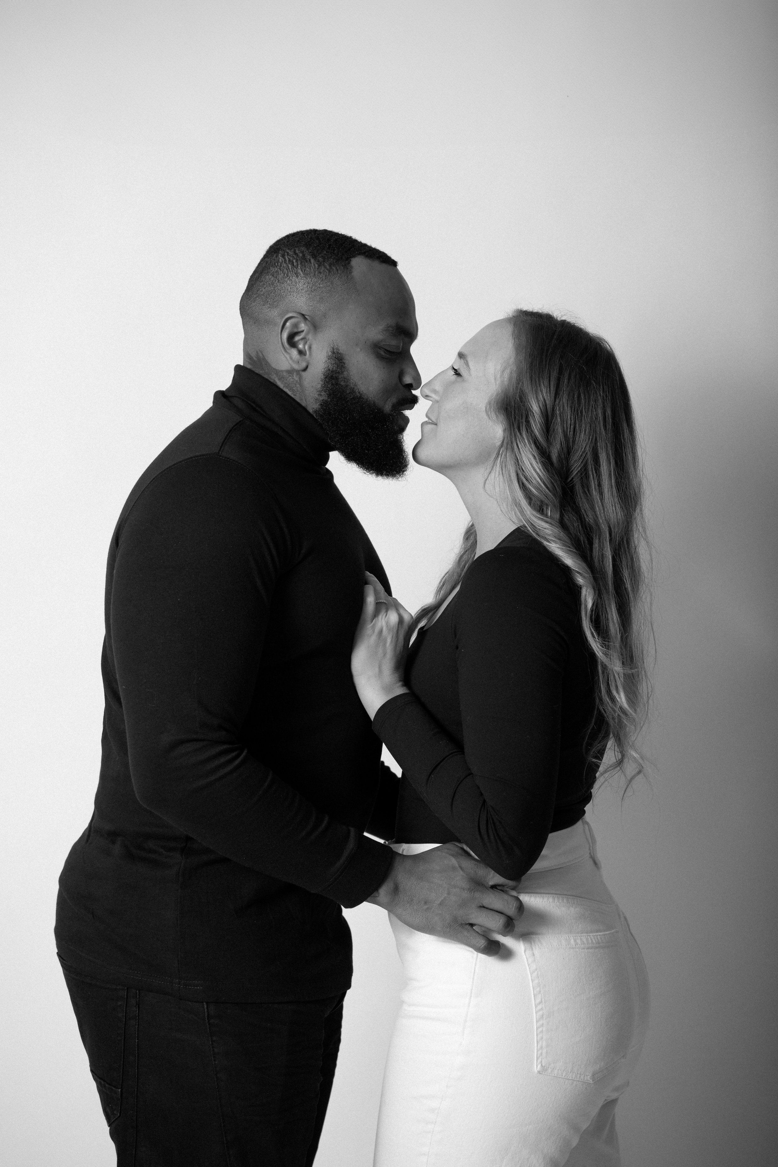 A black-and-white photo of a couple about to kiss, standing close with their noses touching, holding each other affectionately against a plain light background.