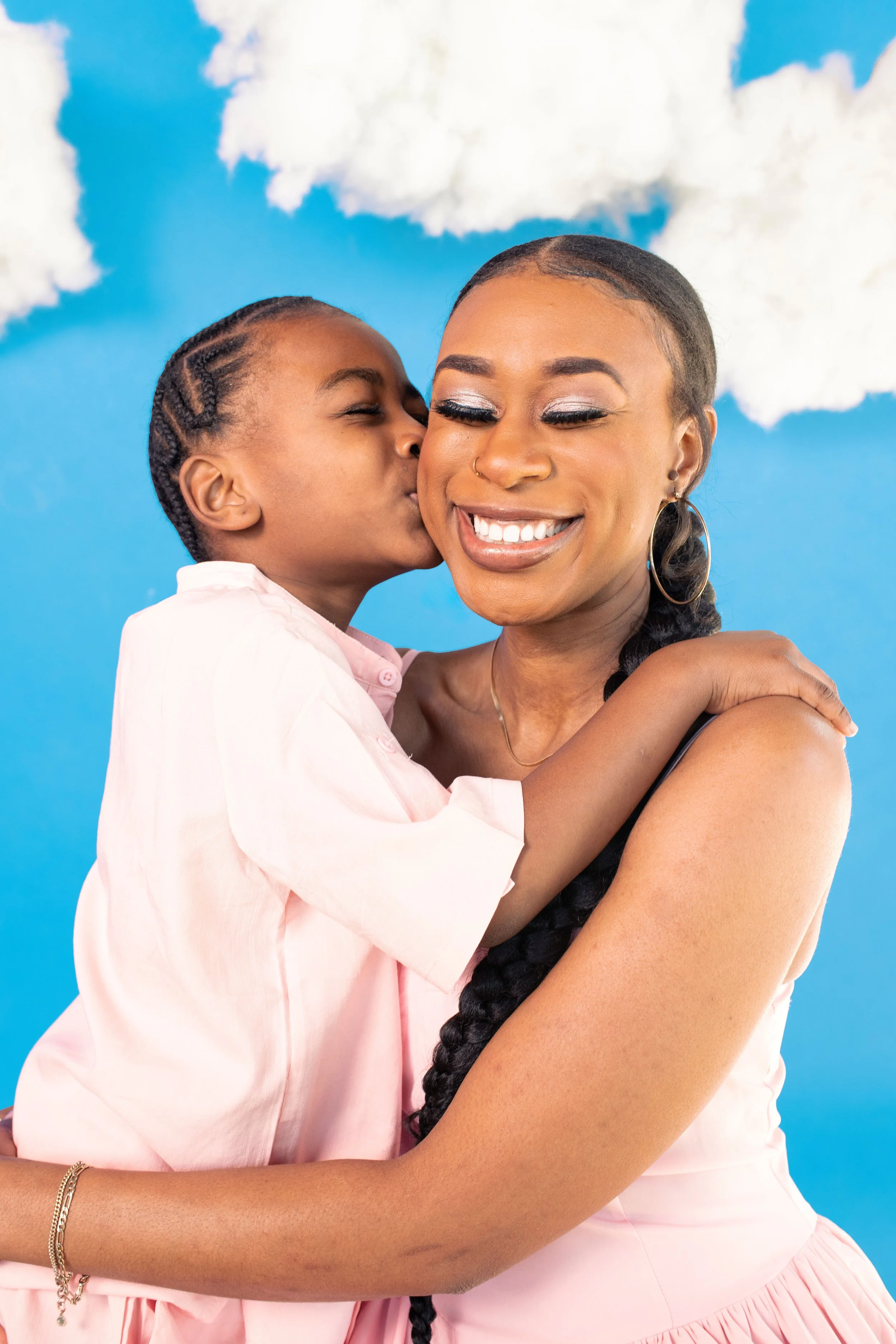 A young girl giving a kiss on the cheek to a smiling woman with long braided hair against a blue sky with clouds background.