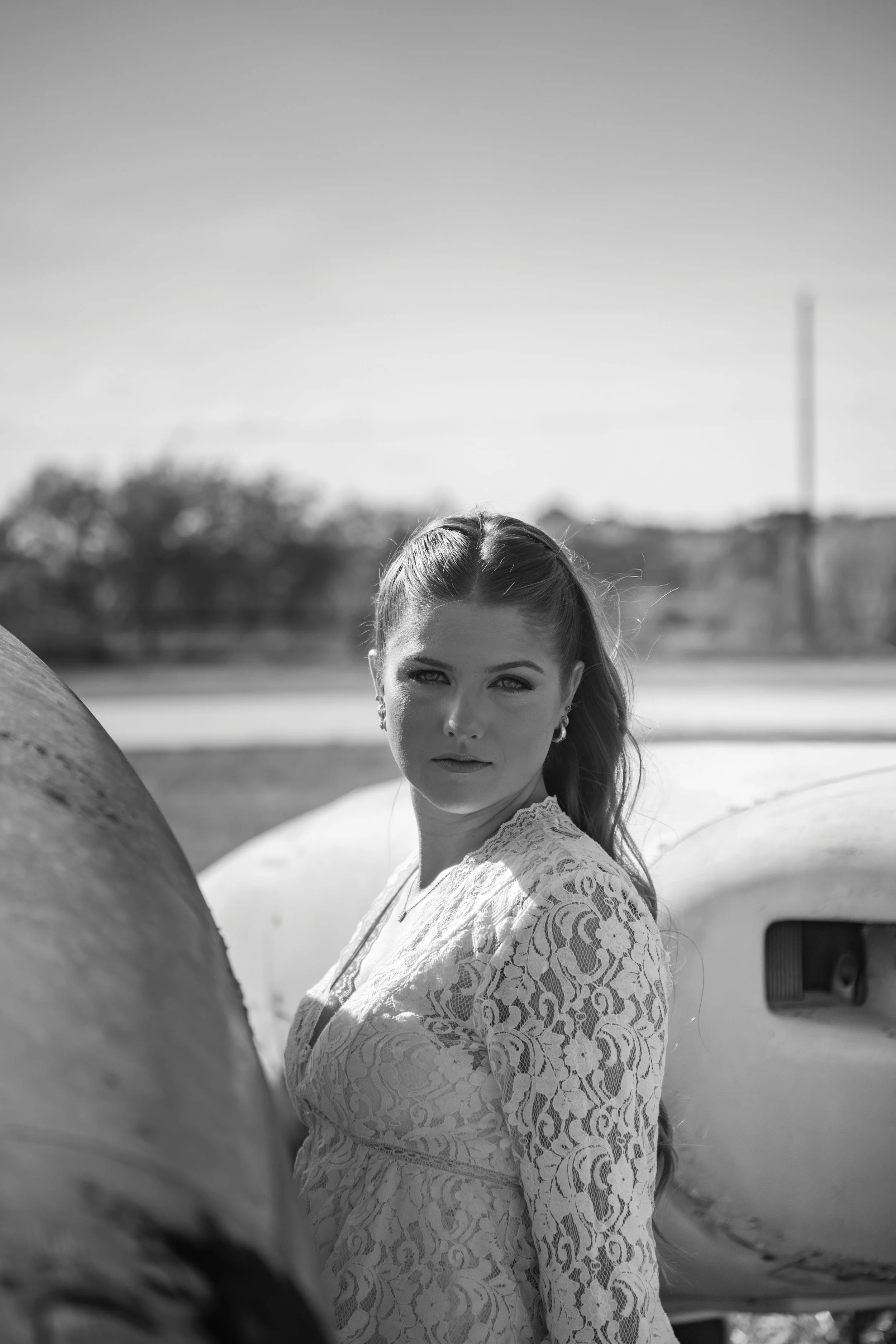 A woman with long hair and patterned lace dress standing outdoors next to a vintage van, with trees in the blurred background, captured in black and white.