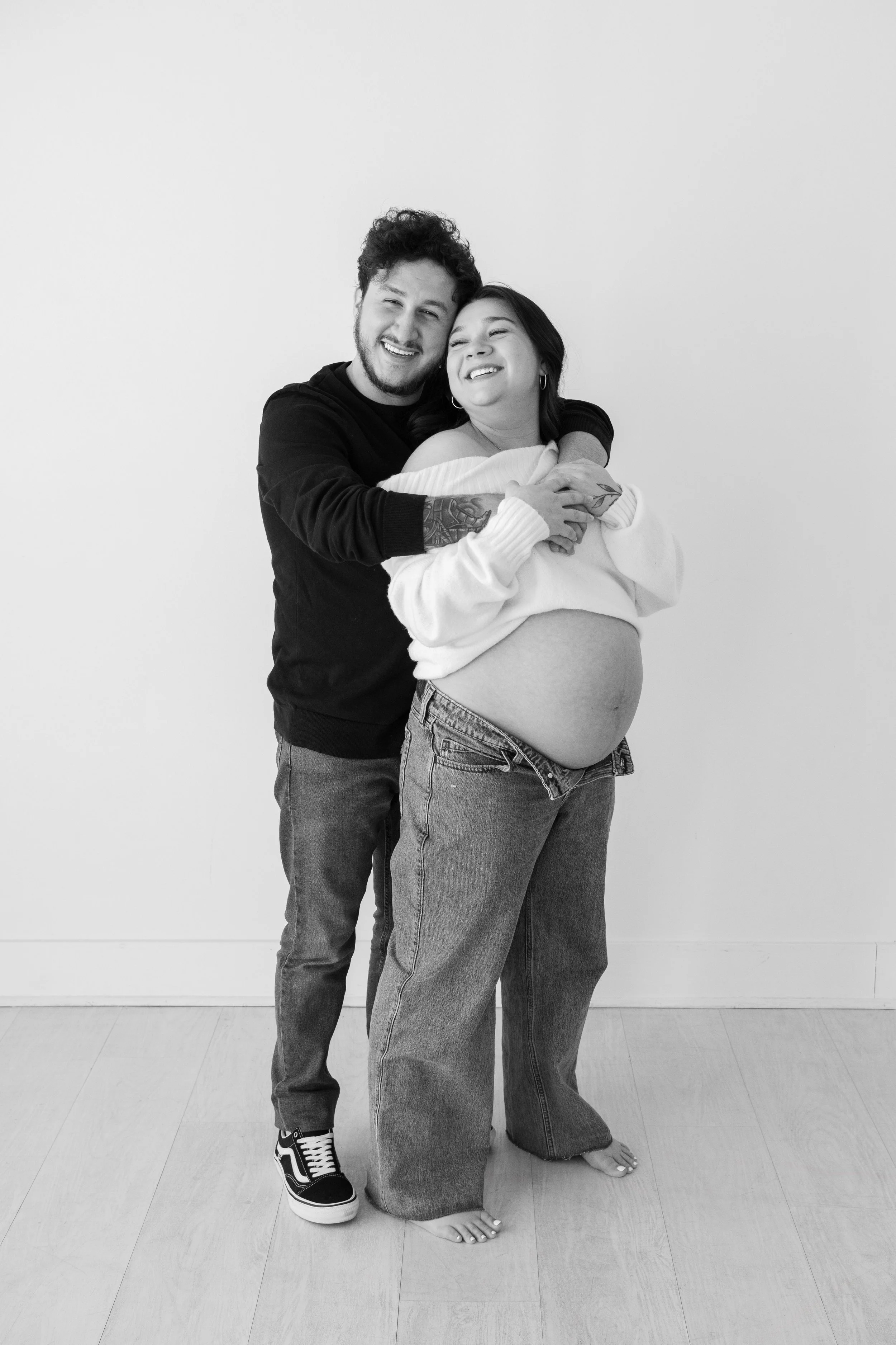 A black and white photo of a smiling couple, with the woman visibly pregnant, embracing each other and standing barefoot on a wooden floor against a plain white wall.