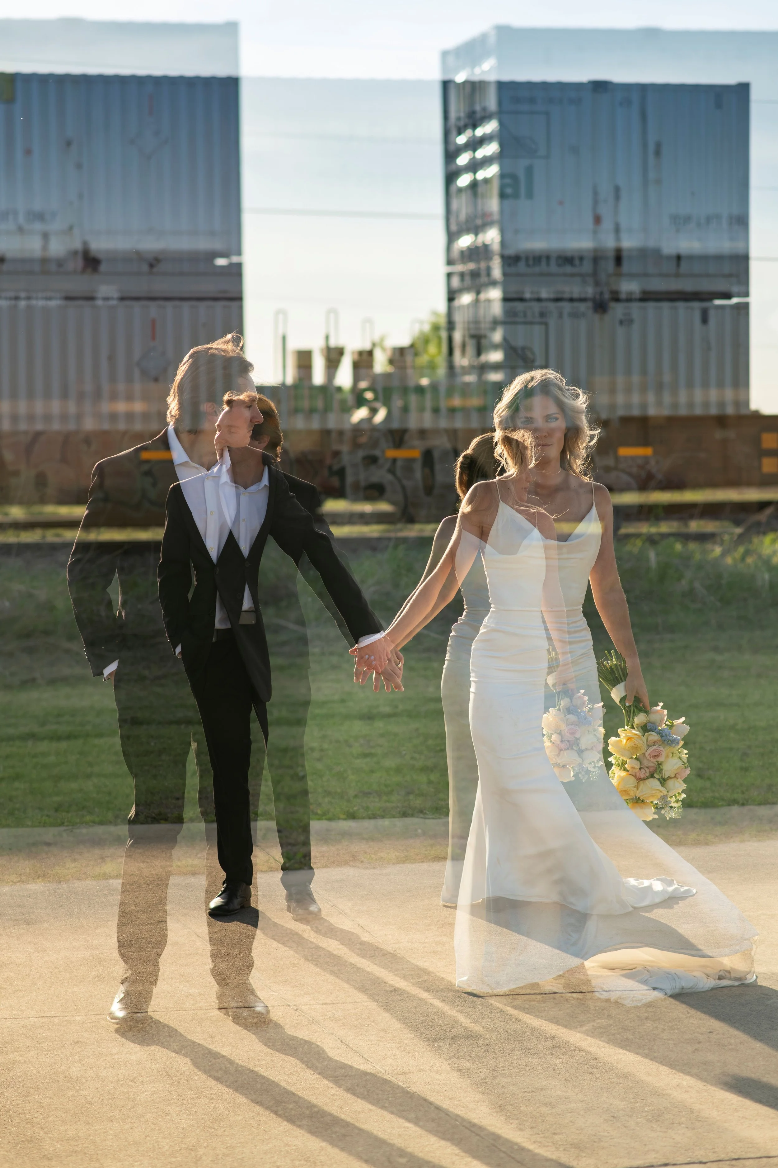 A bride and groom holding hands and walking outside, with a reflection of their images overlapping. The bride is in a white wedding dress holding a bouquet, and the groom is in a black suit. There are blue sky and industrial buildings in the backgrou