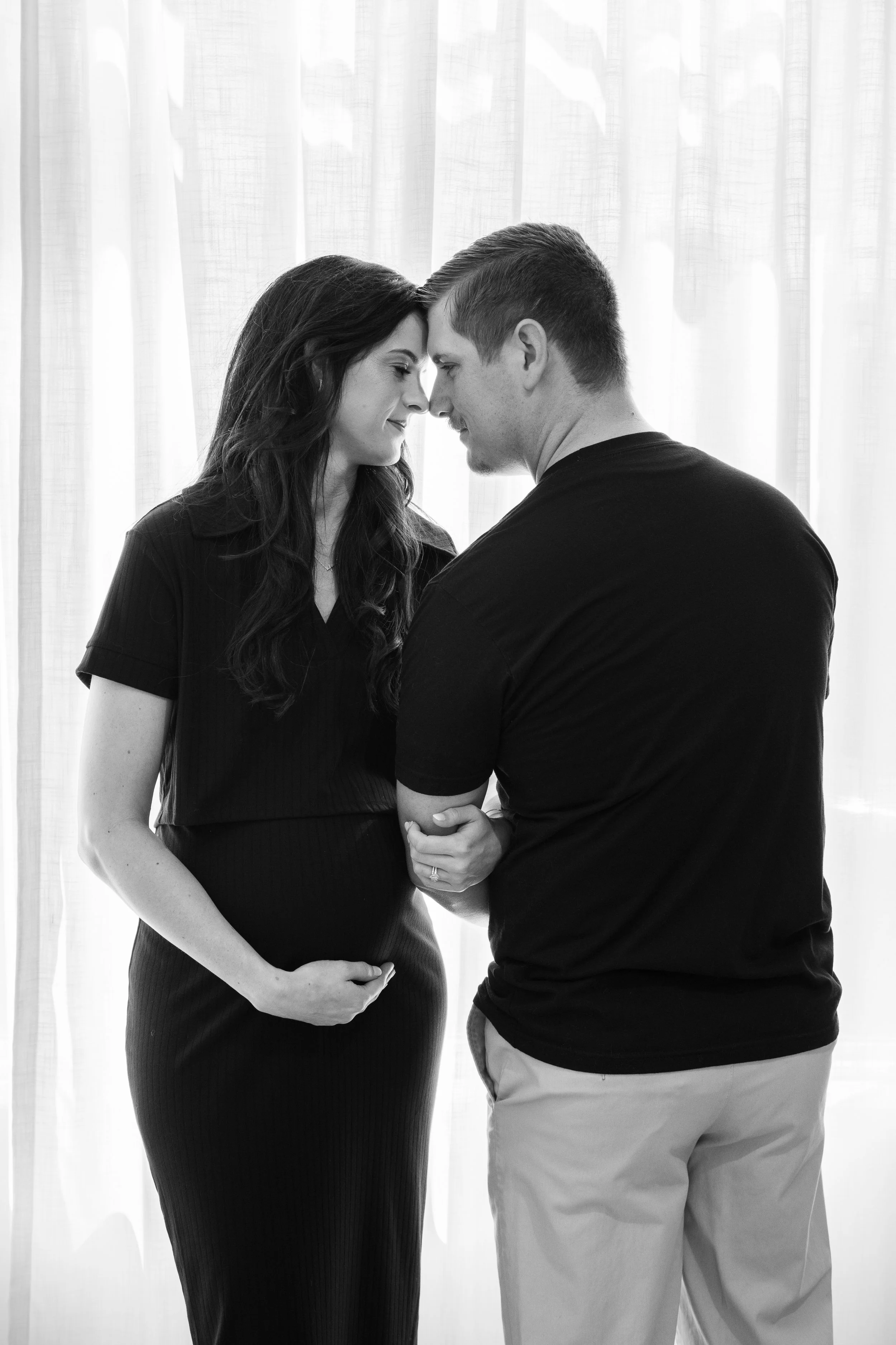 A couple facing each other with foreheads touching, the woman is pregnant and gently holding her belly, both smiling softly.