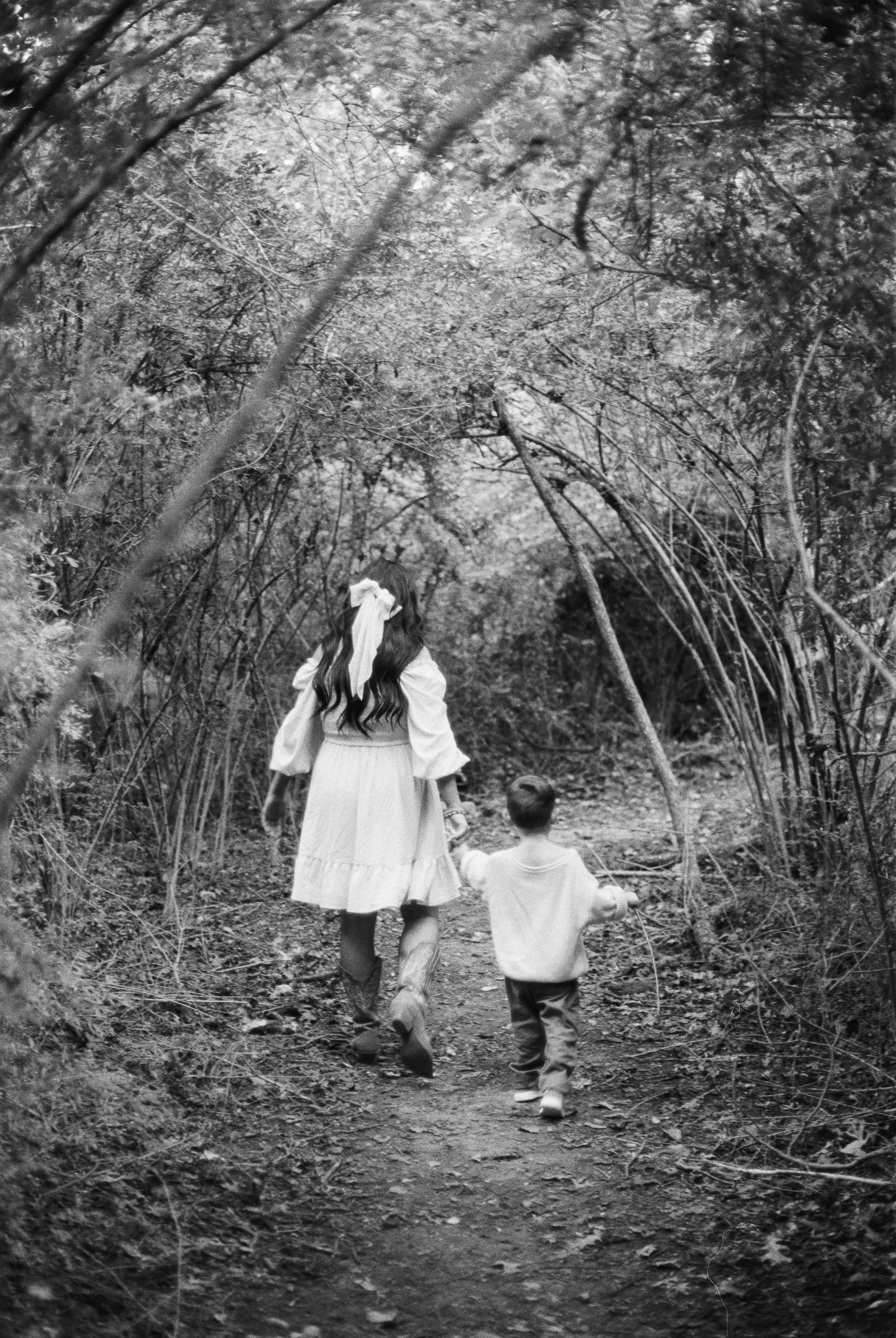 A woman and a young boy walk hand in hand on a forest trail surrounded by dense trees and foliage in black and white.