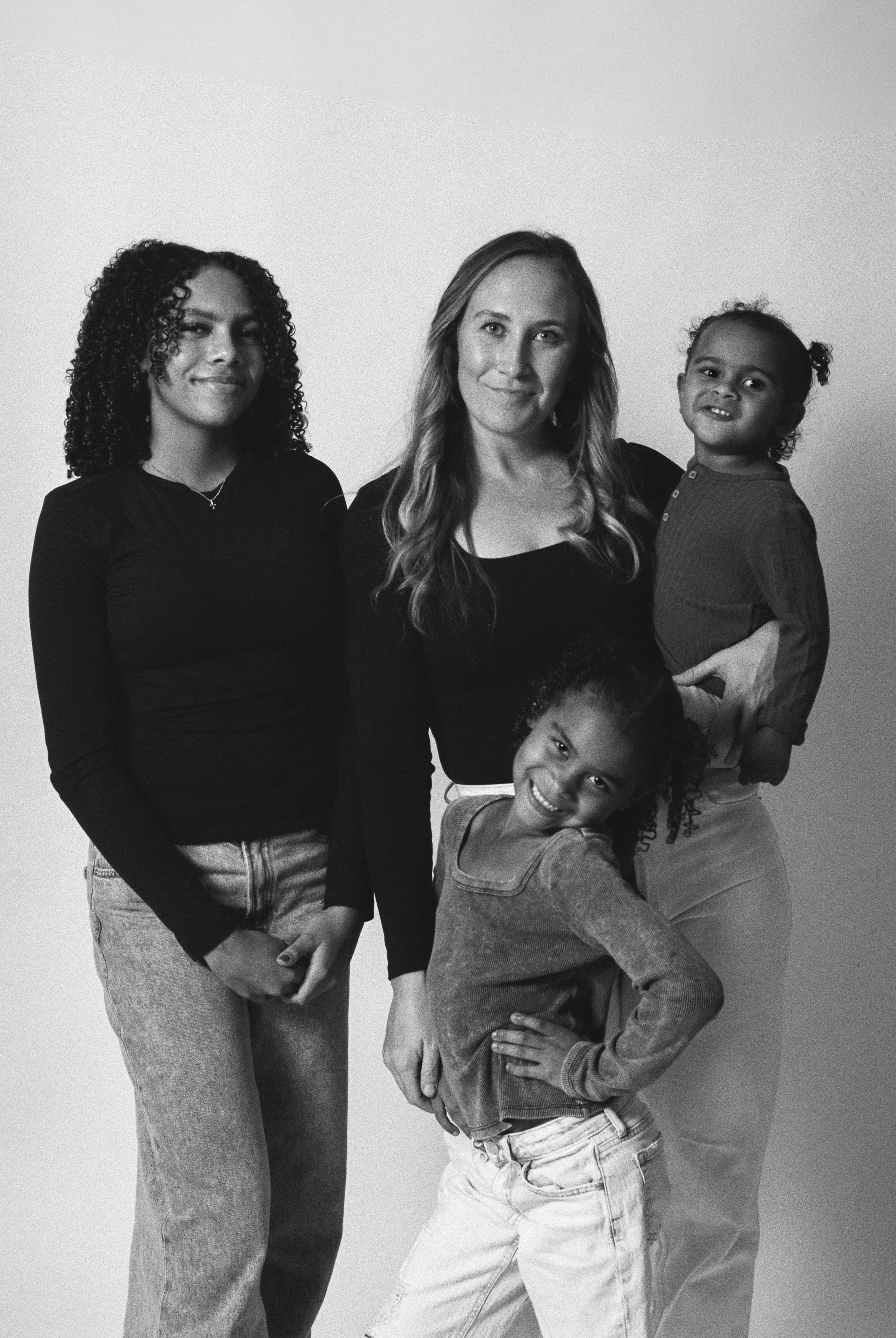 Black and white photo of a woman with three young girls, all smiling and dressed casually, standing against a plain background.