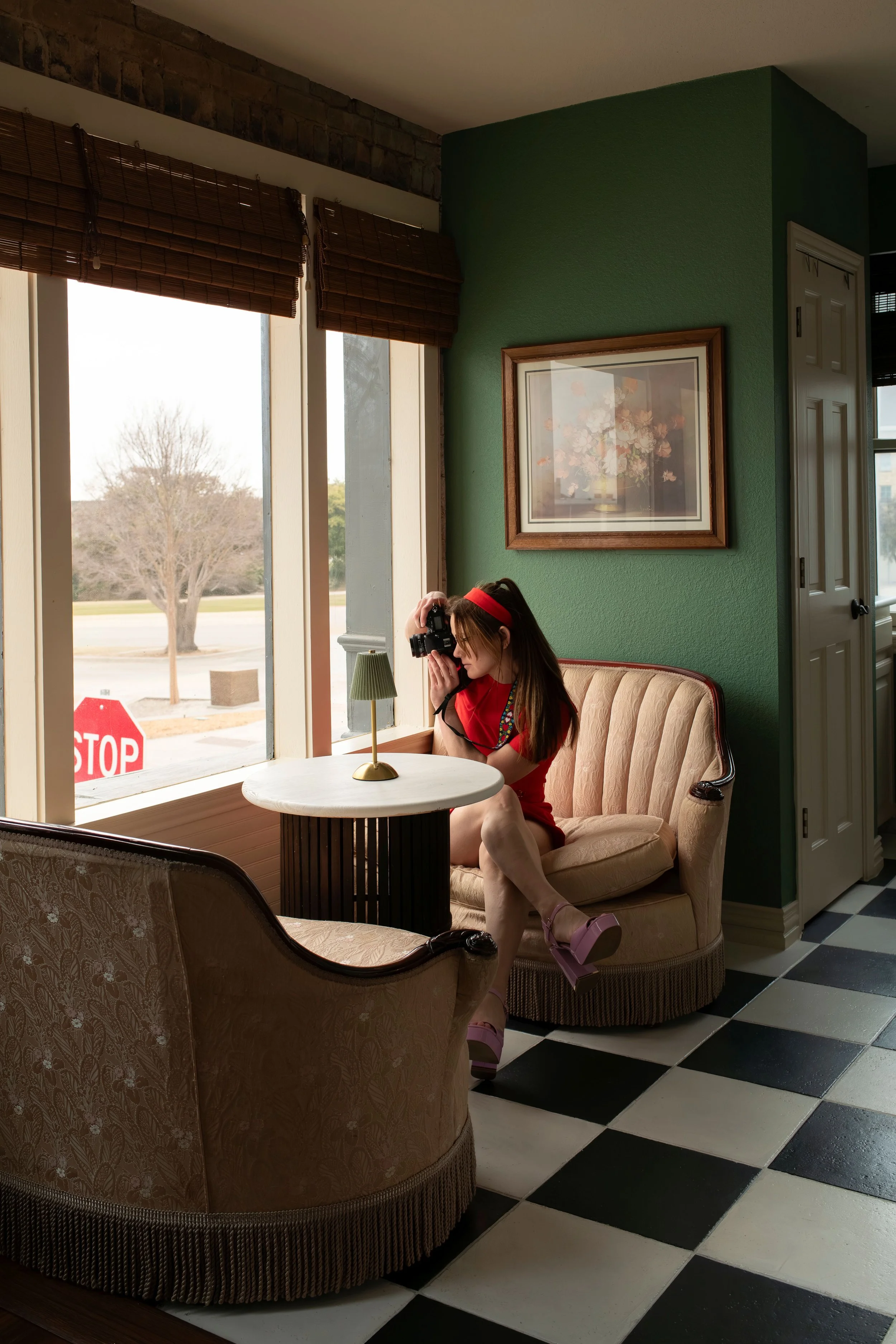 A woman dressed in a red dress and pink high heels sitting in a vintage booth with a camera up to her face, near a window with a view of a street, trees, a stop sign, and an outdoor scene.