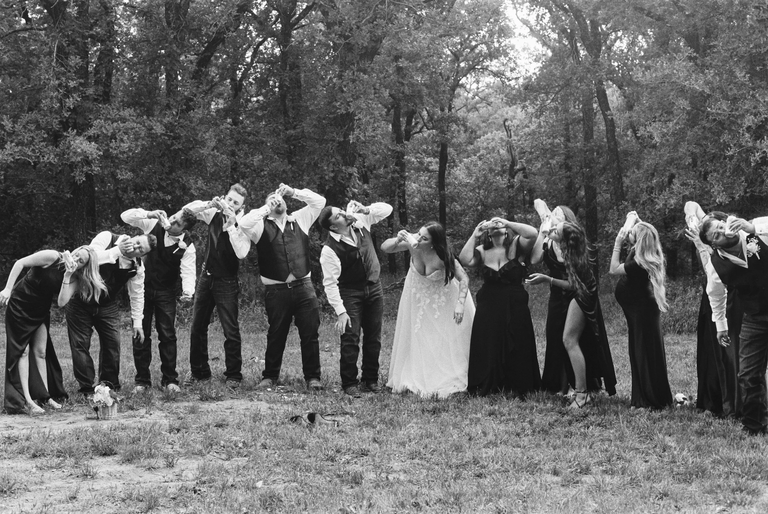 A group of people, including men and women, standing outdoors in a line and drinking from cans, with many trees in the background. The photo is in black and white.