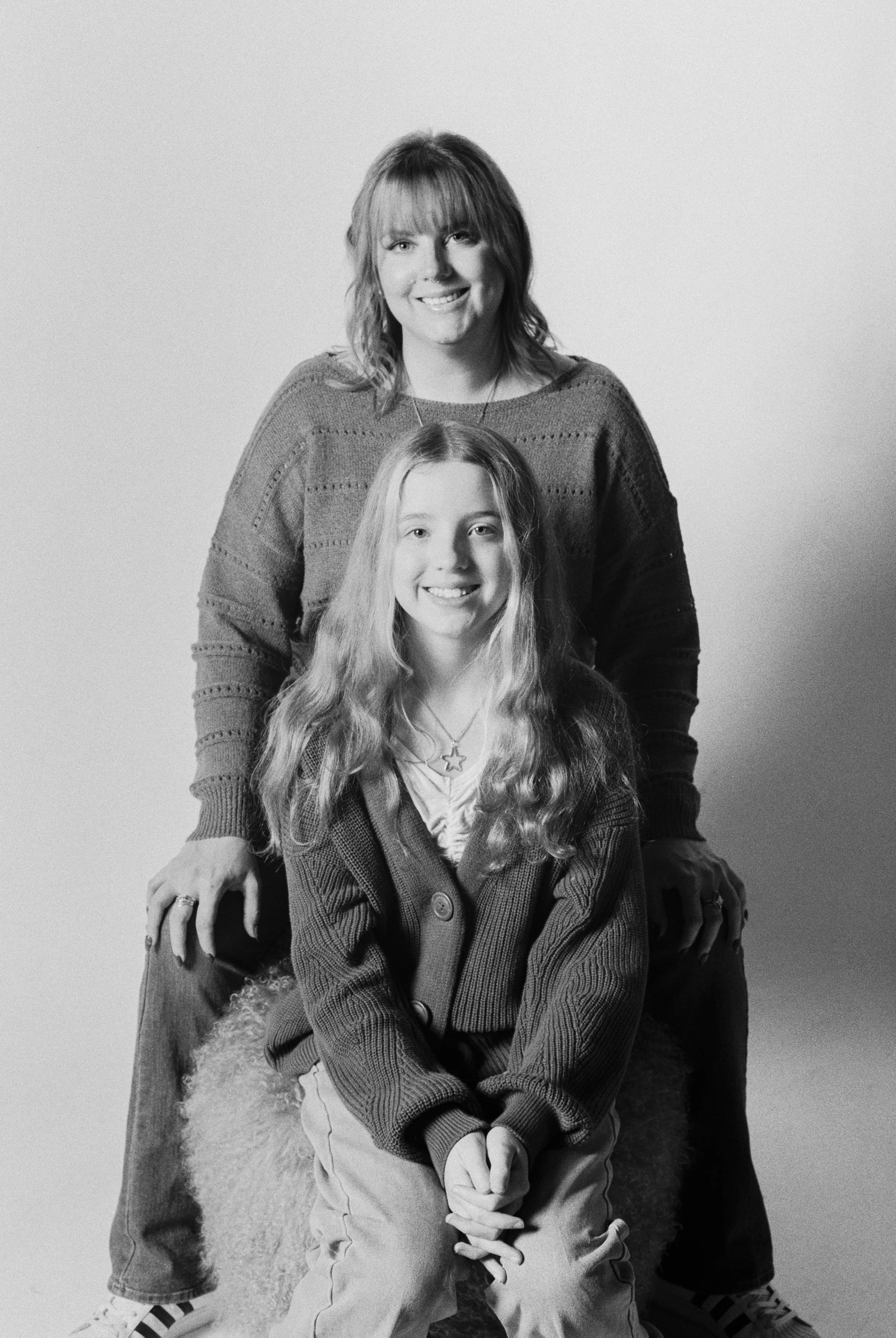 Black and white photo of a woman and a girl, both smiling. The woman stands behind the girl, who is sitting on a fluffy stool, and they both wear sweaters.