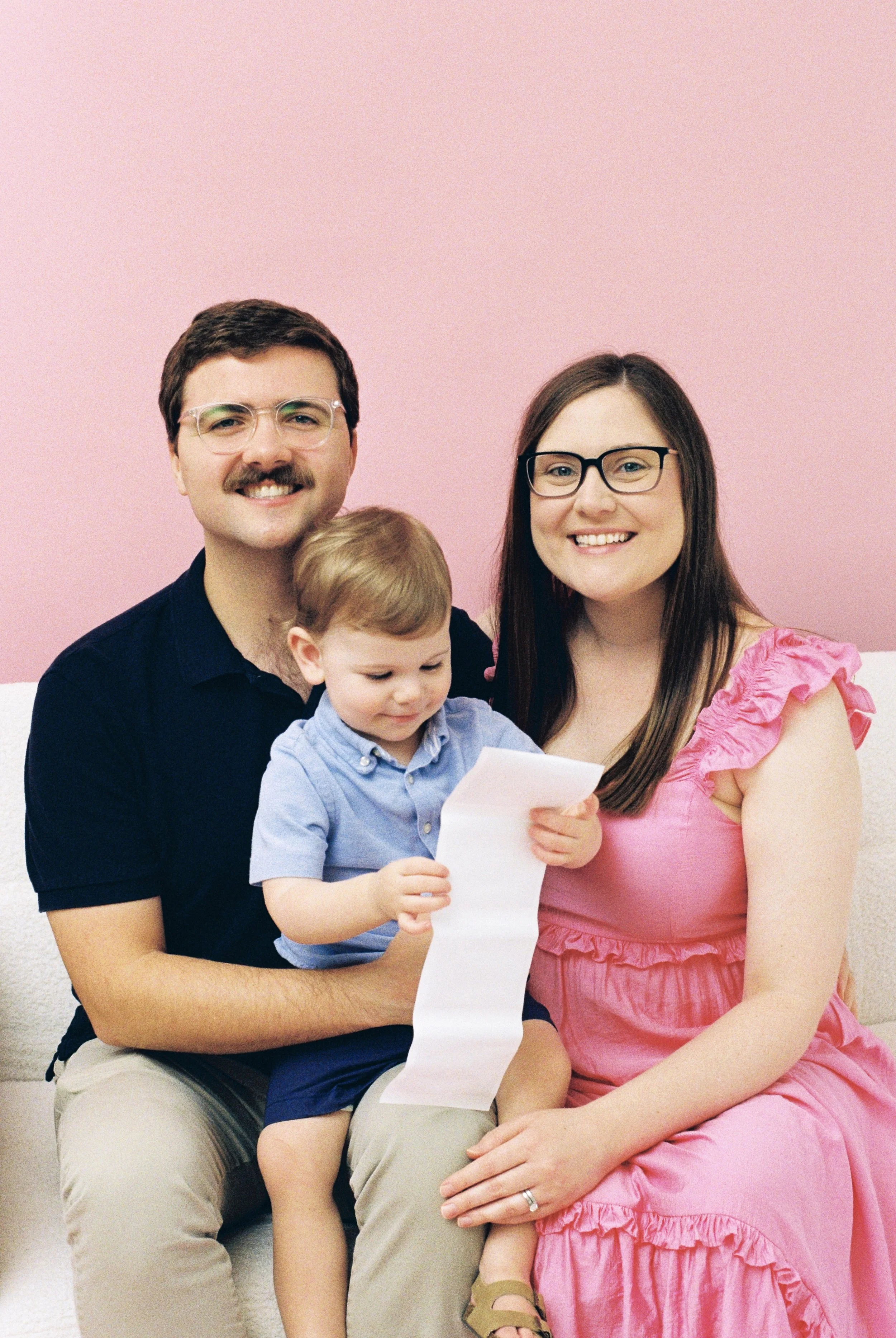 A family of three: a man, woman, and young boy, sitting on a white couch against a pink wall. The boy is looking at a long piece of paper, with the man and woman smiling.
