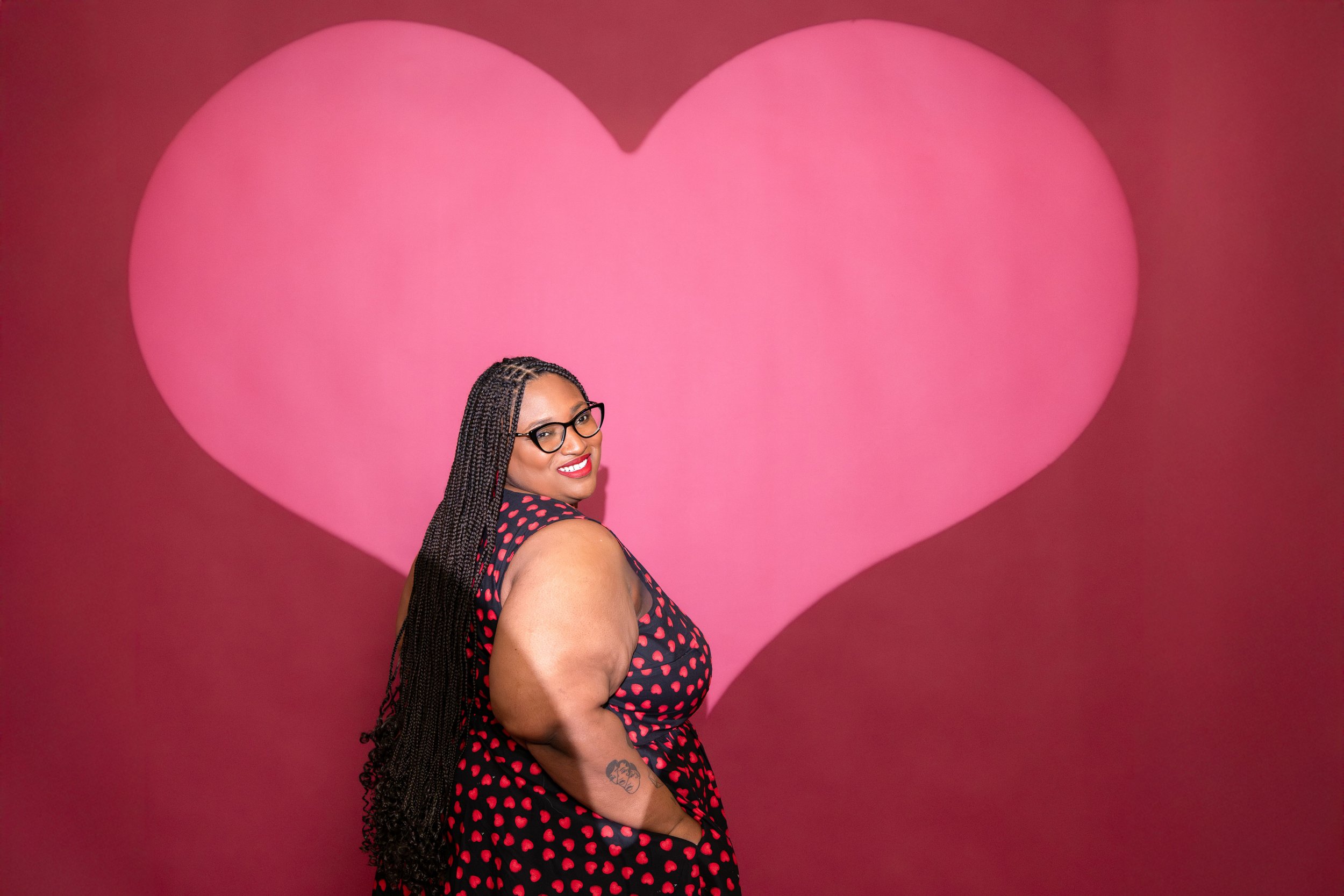 A woman with long braided hair, glasses, wearing a black dress with pink heart pattern, smiling, standing in front of a large pink heart background.