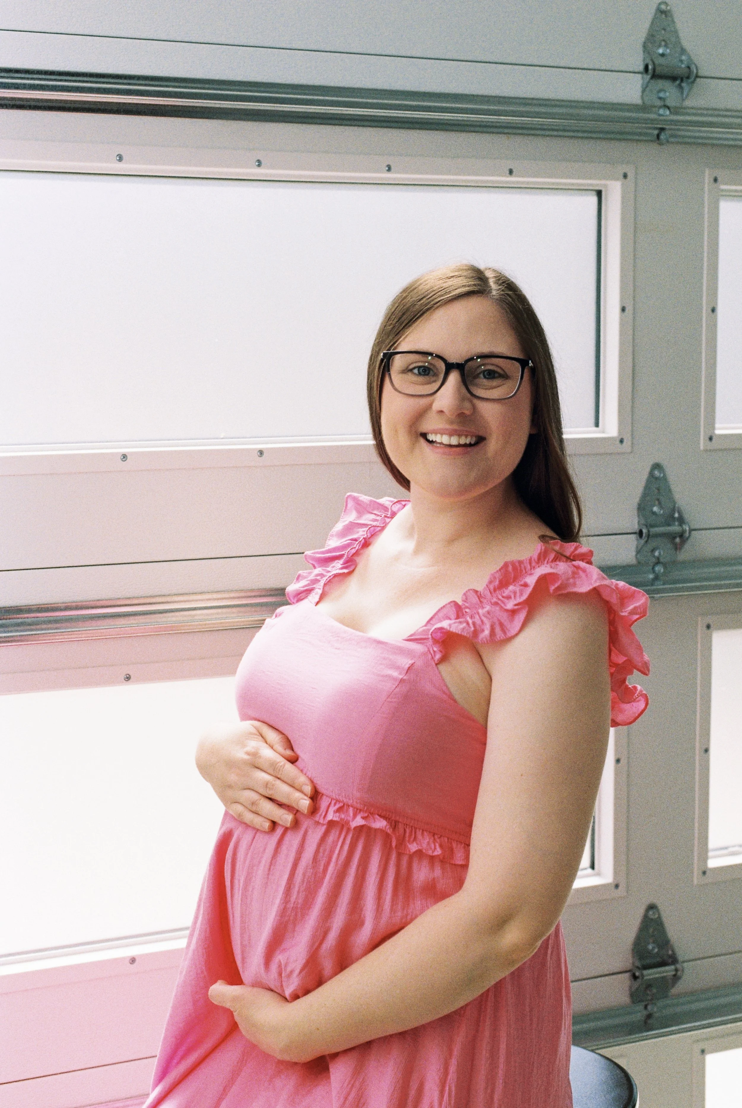A smiling pregnant woman with glasses, wearing a pink dress with ruffled sleeves, standing in front of a garage door.
