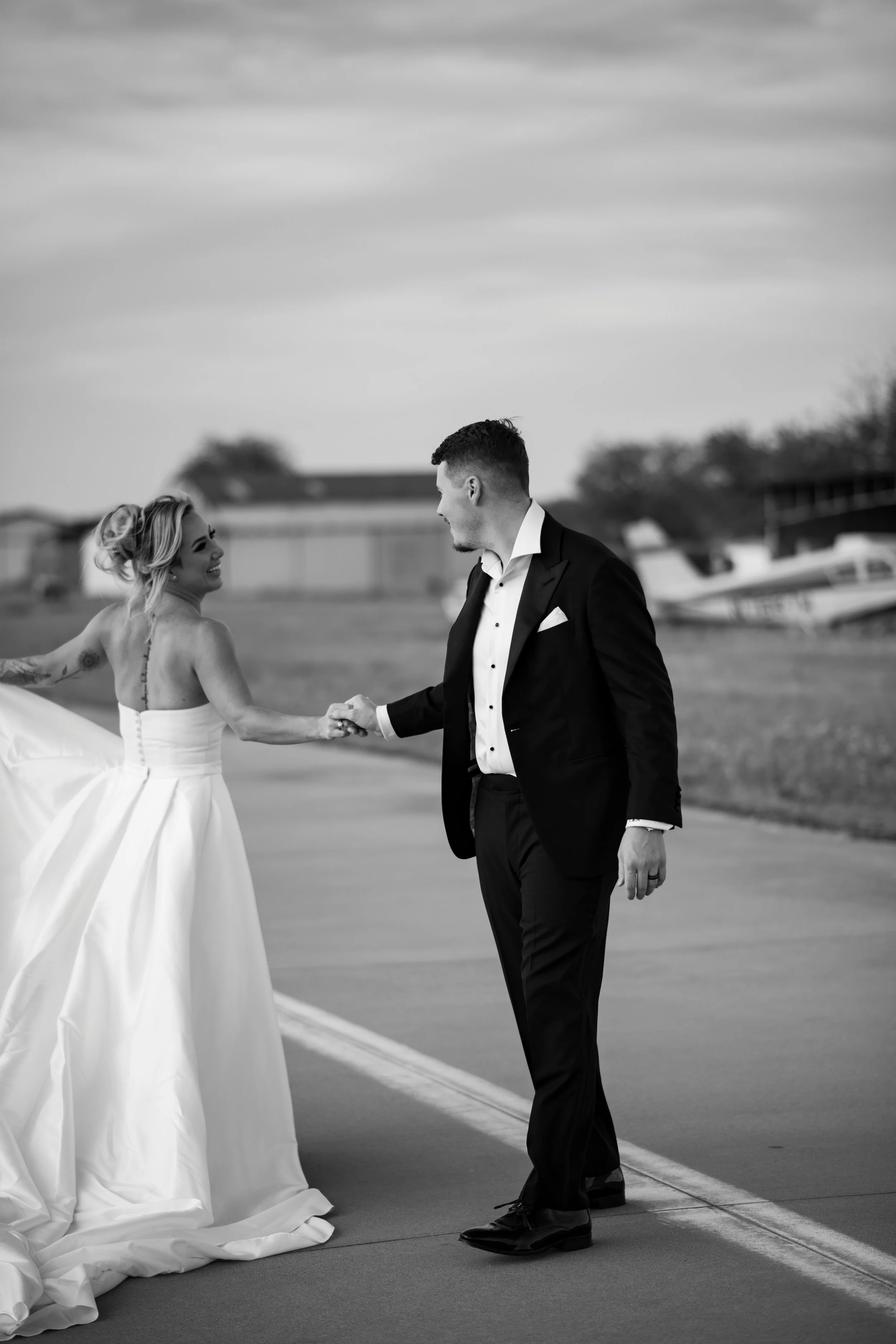 A black and white photo of a newlywed couple at an airport, holding hands and smiling at each other. The bride is in a strapless gown with her hair in an updo, and the groom is in a black tuxedo. The background features small airplanes parked on the 