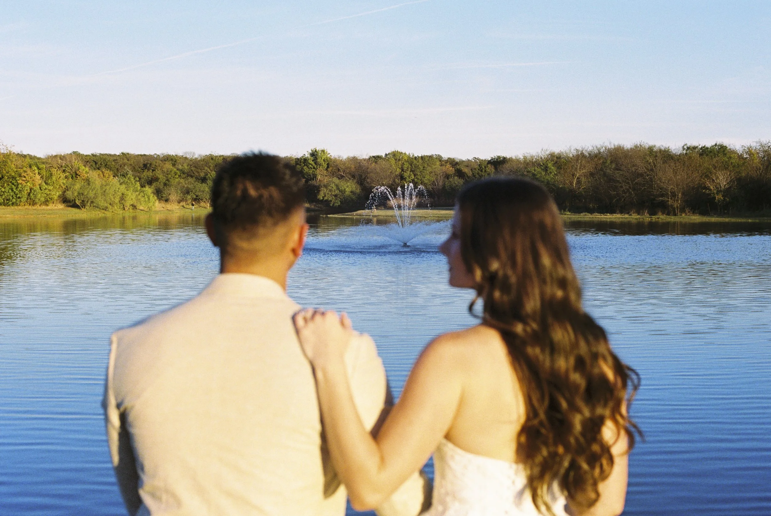 A couple by a lake with a fountain, surrounded by trees, during sunset.