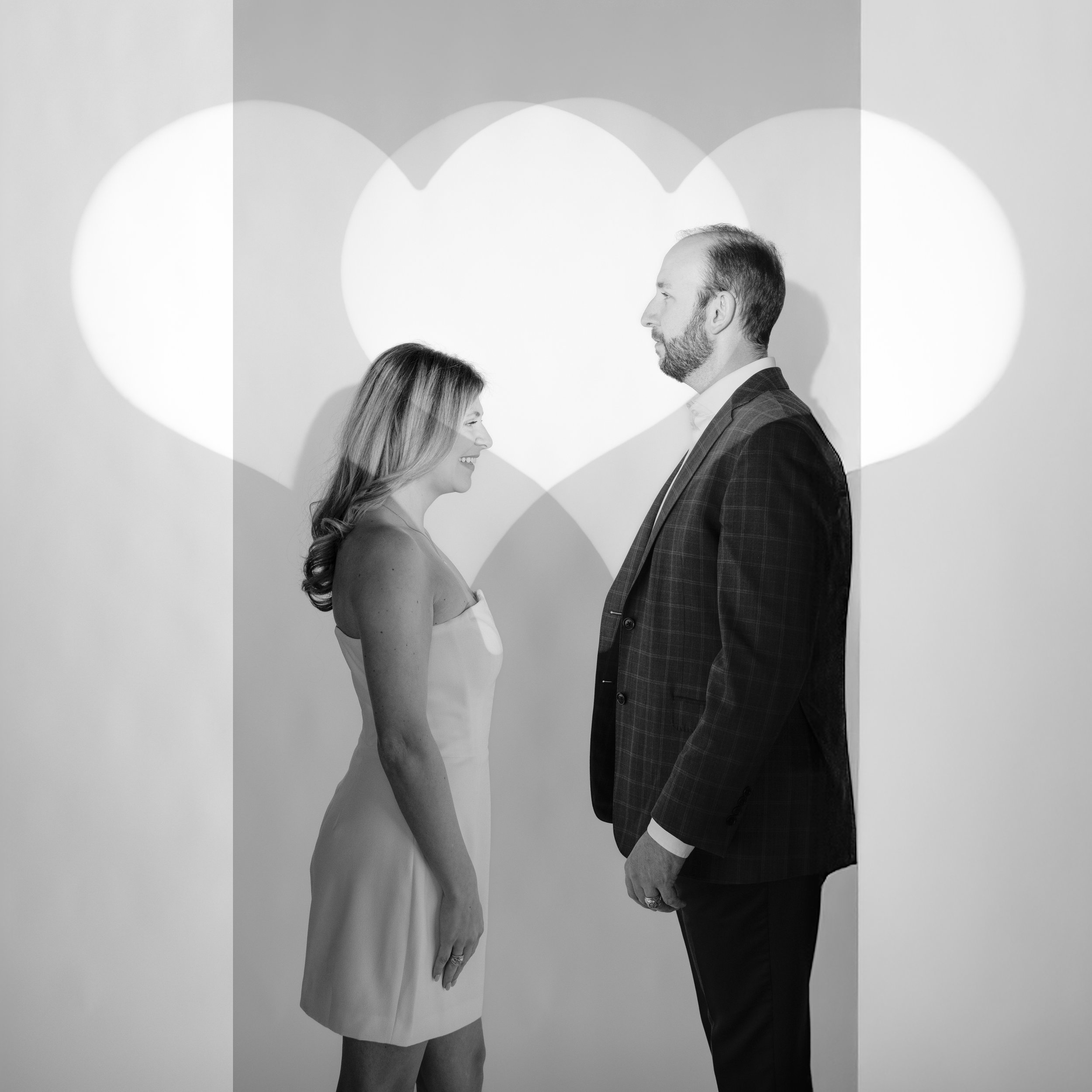 A black and white photo of a woman and a man facing each other, smiling, with large heart-shaped light projections in the background.