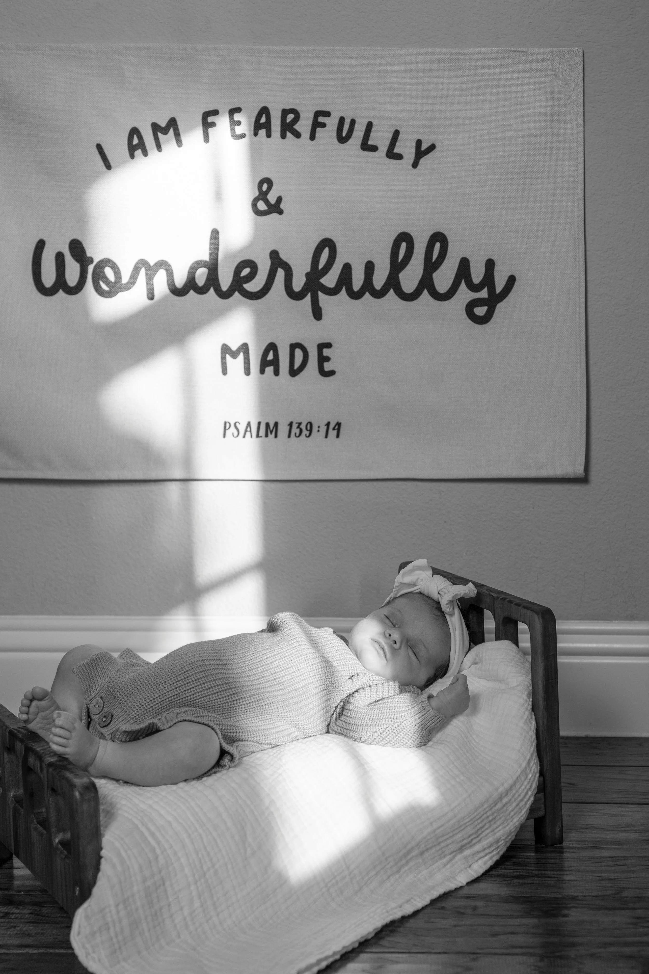A sleeping baby lying on a small bed or cradle, wearing a knitted outfit and a headband, next to a wall with a biblical quote on it in a room with hardwood floor.