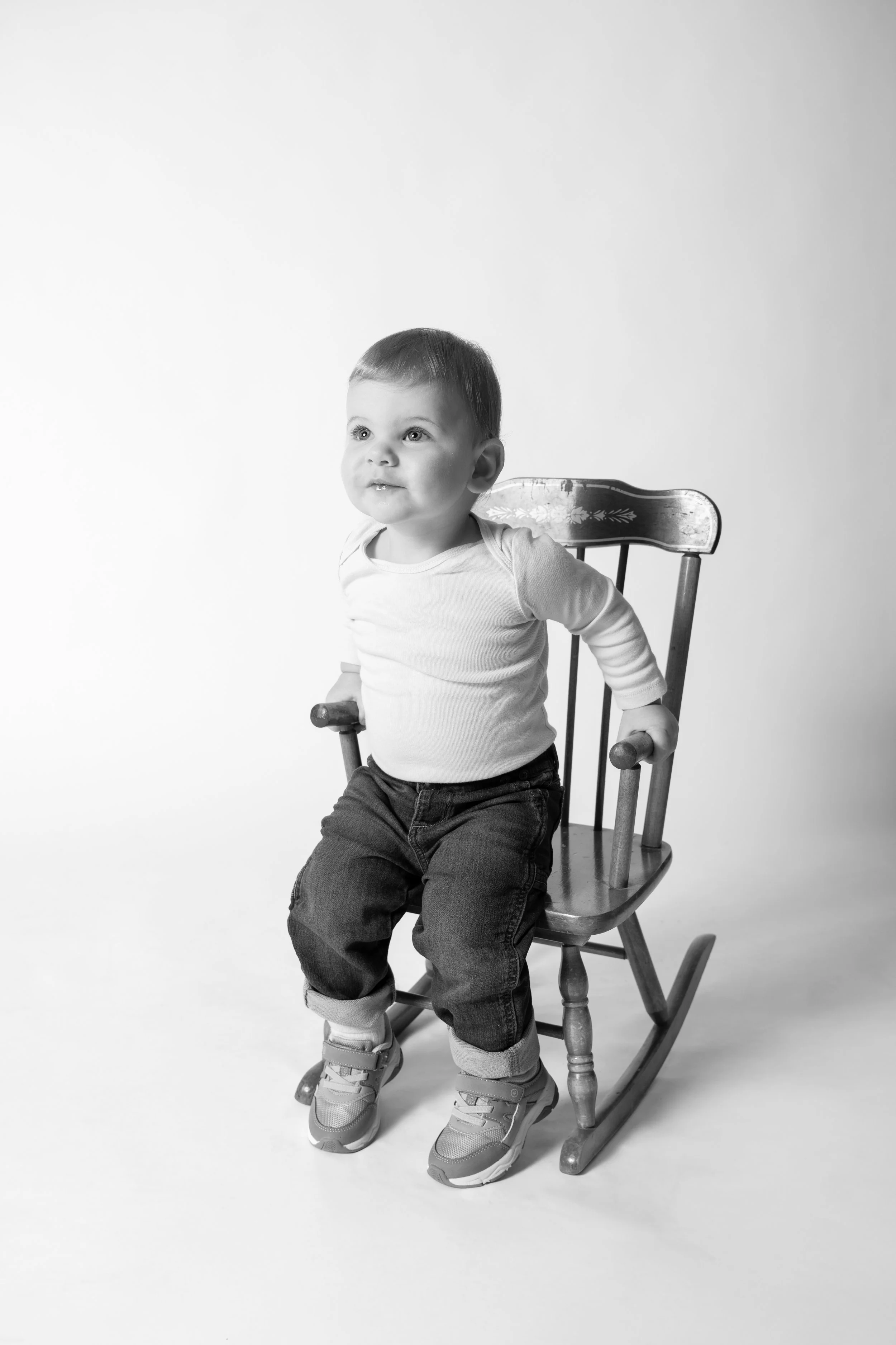 Black and white photo of a young child sitting on a wooden rocking chair, looking slightly upward with a curious expression.