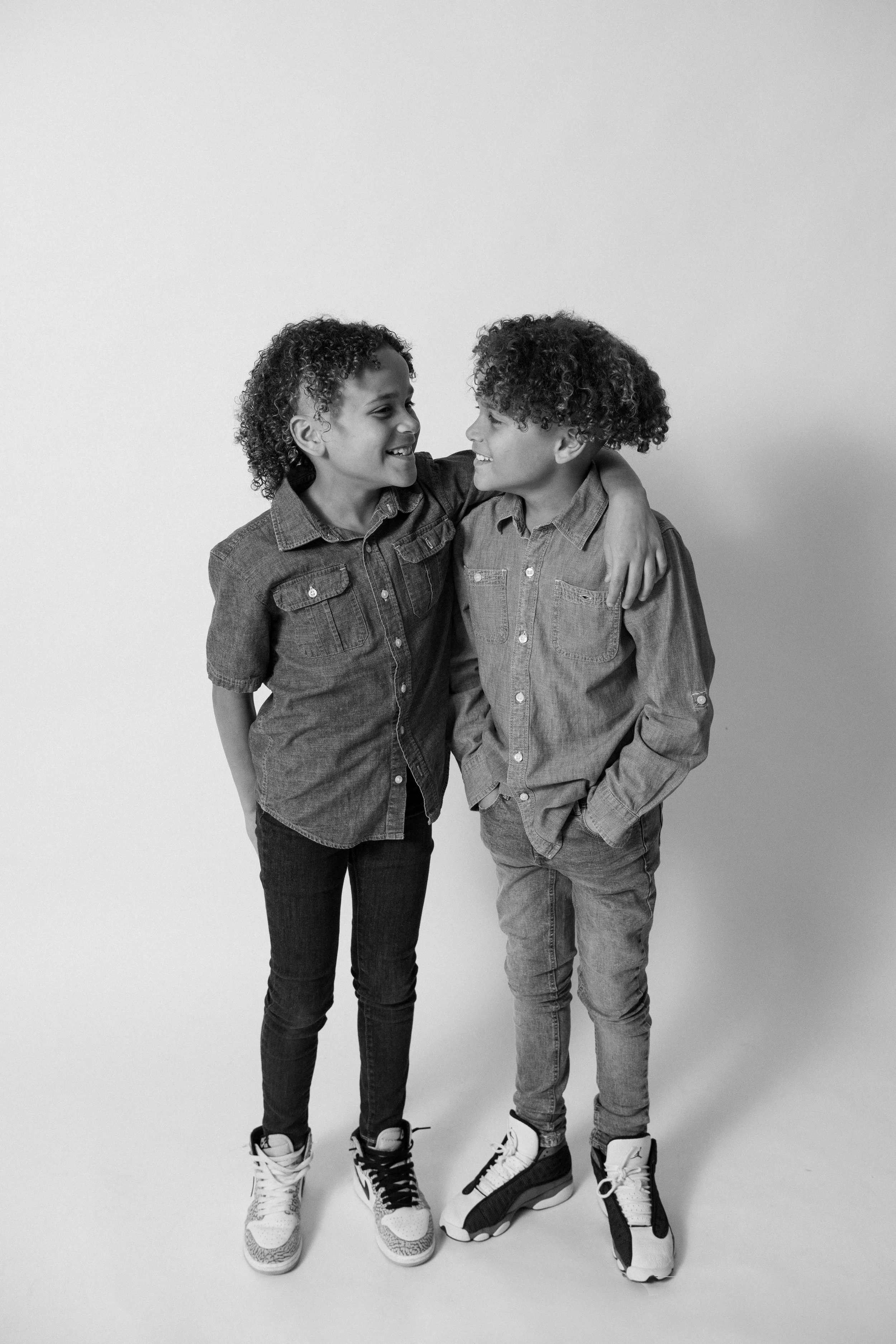 Two young boys with curly hair, wearing denim shirts and sneakers, smiling and interacting affectionately.