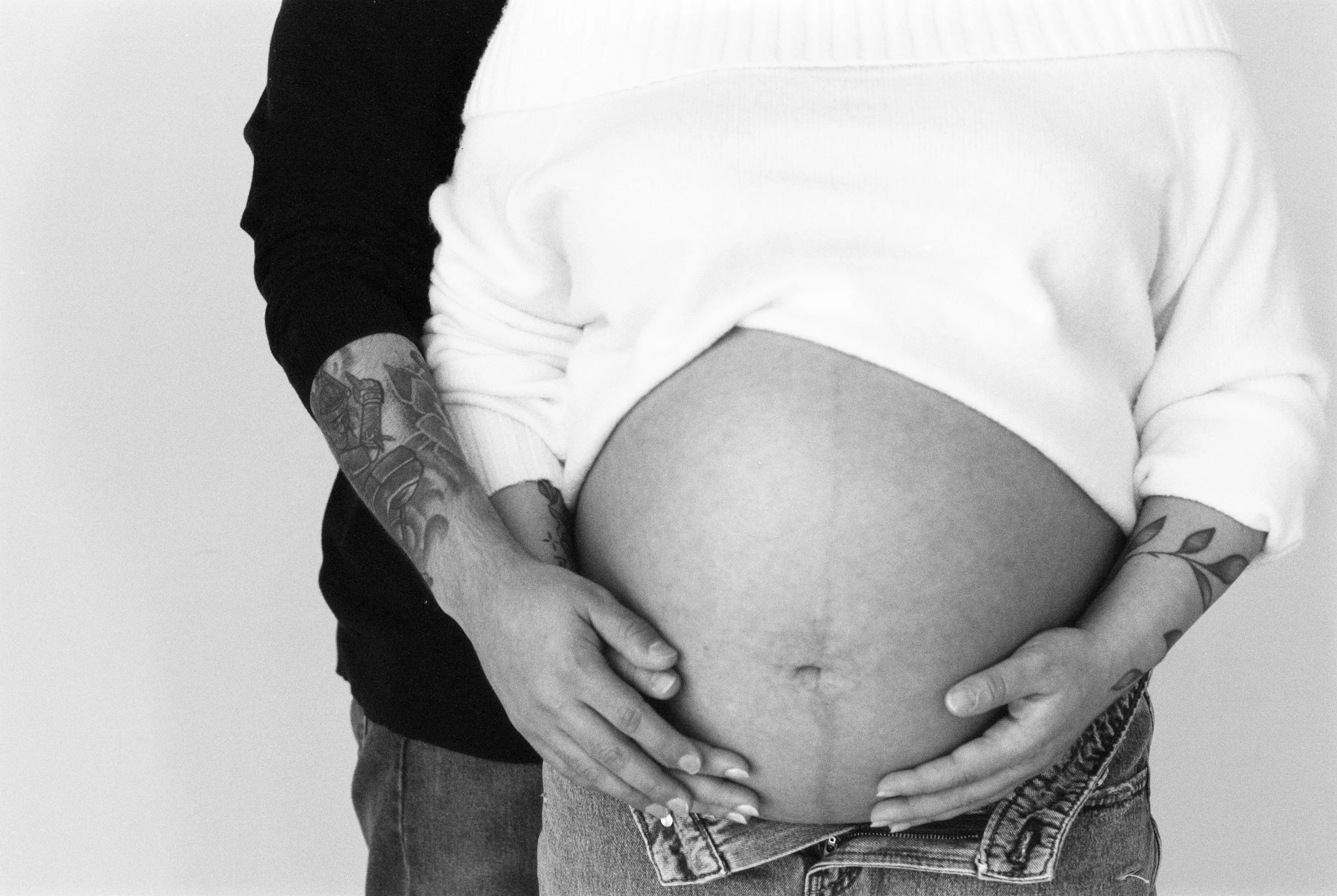 A pregnant woman with tattoos on her arms, wearing a white top, is playing with her belly while a man in a black and white shirt stands behind her, also touching her belly.