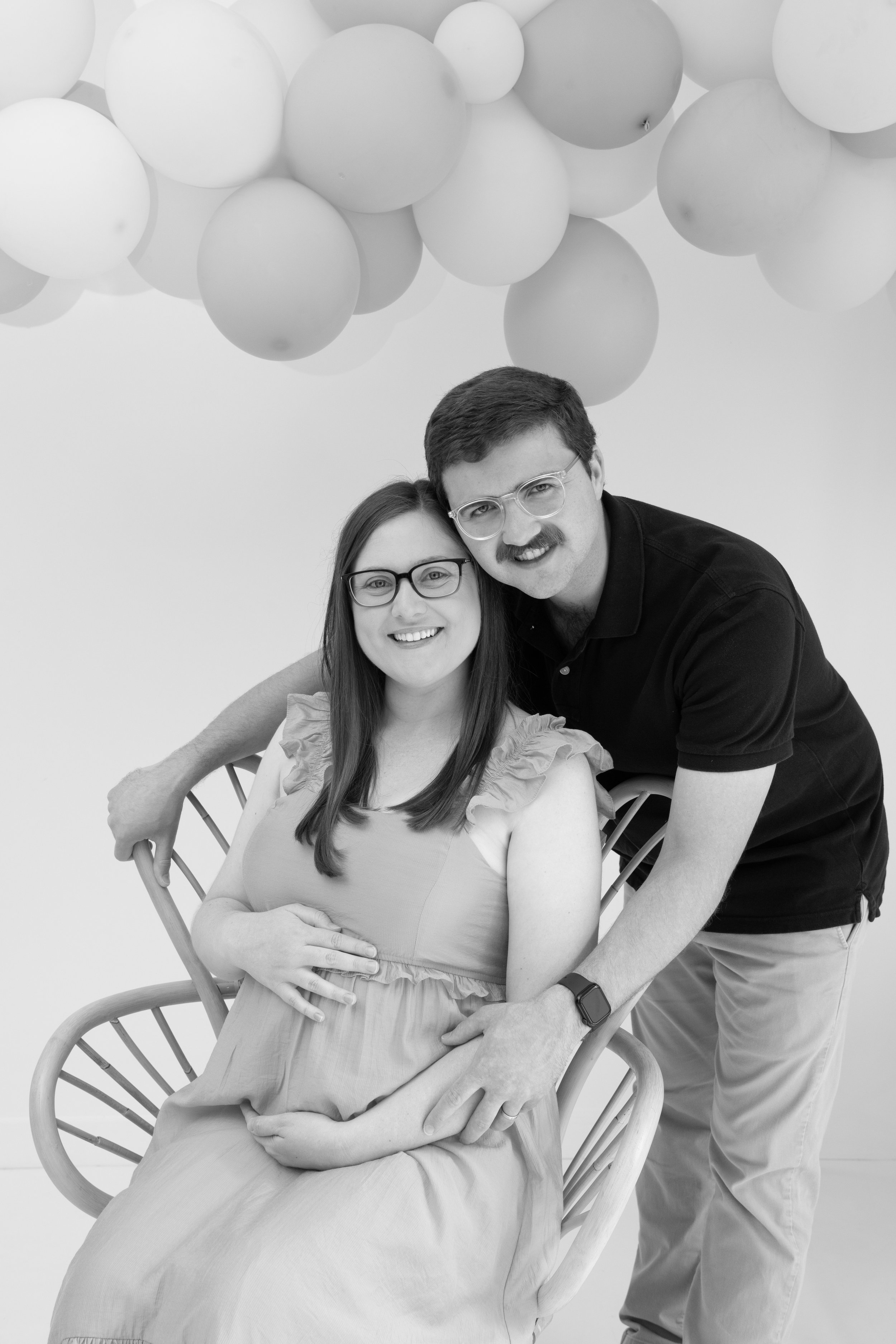A pregnant woman sitting on a modern wooden chair, smiling, with a man standing beside her, both wearing glasses, in front of a wall with hanging balloons.