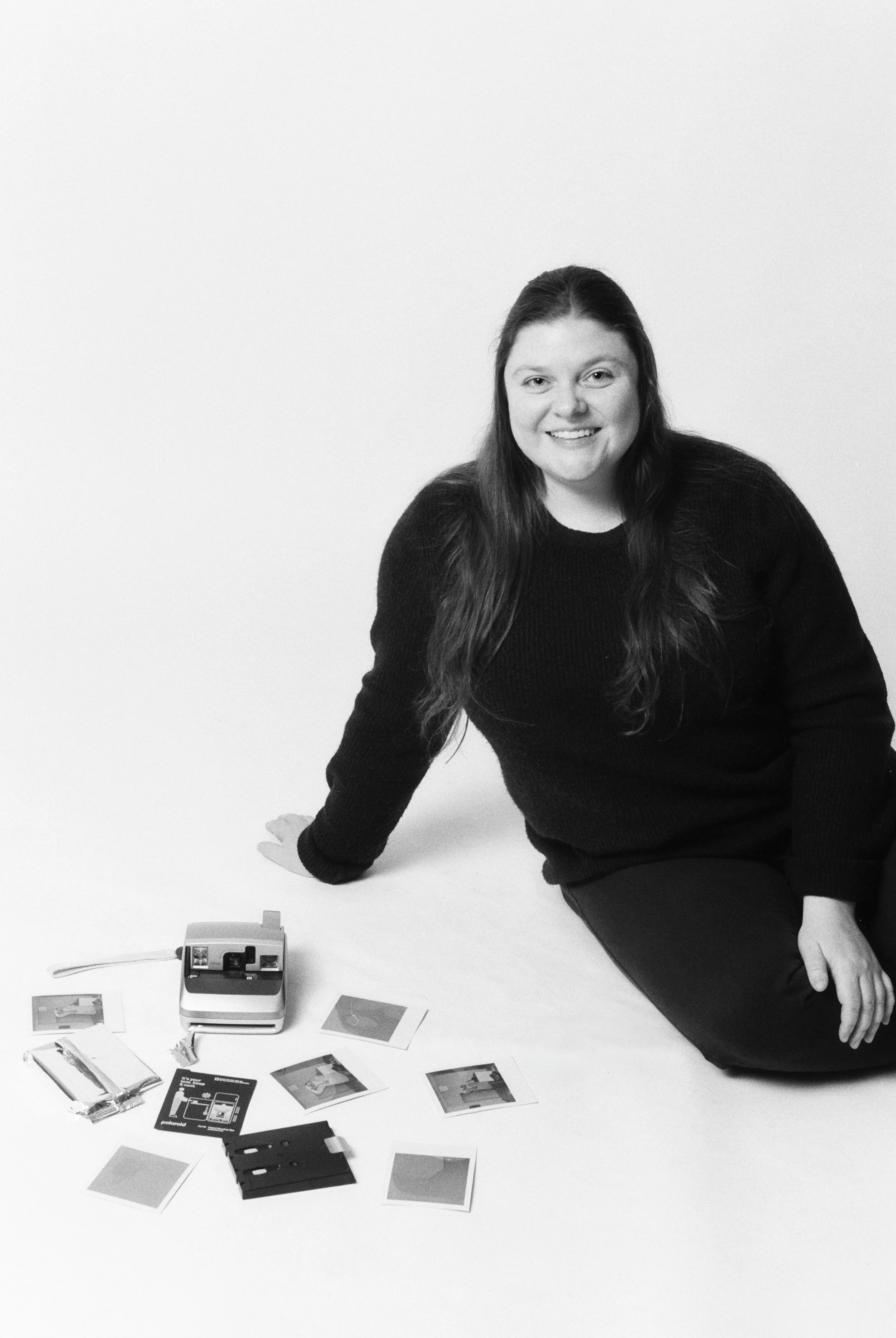 A woman with long hair, smiling, sitting on the floor in front of a camera and various polaroids on a plain background.