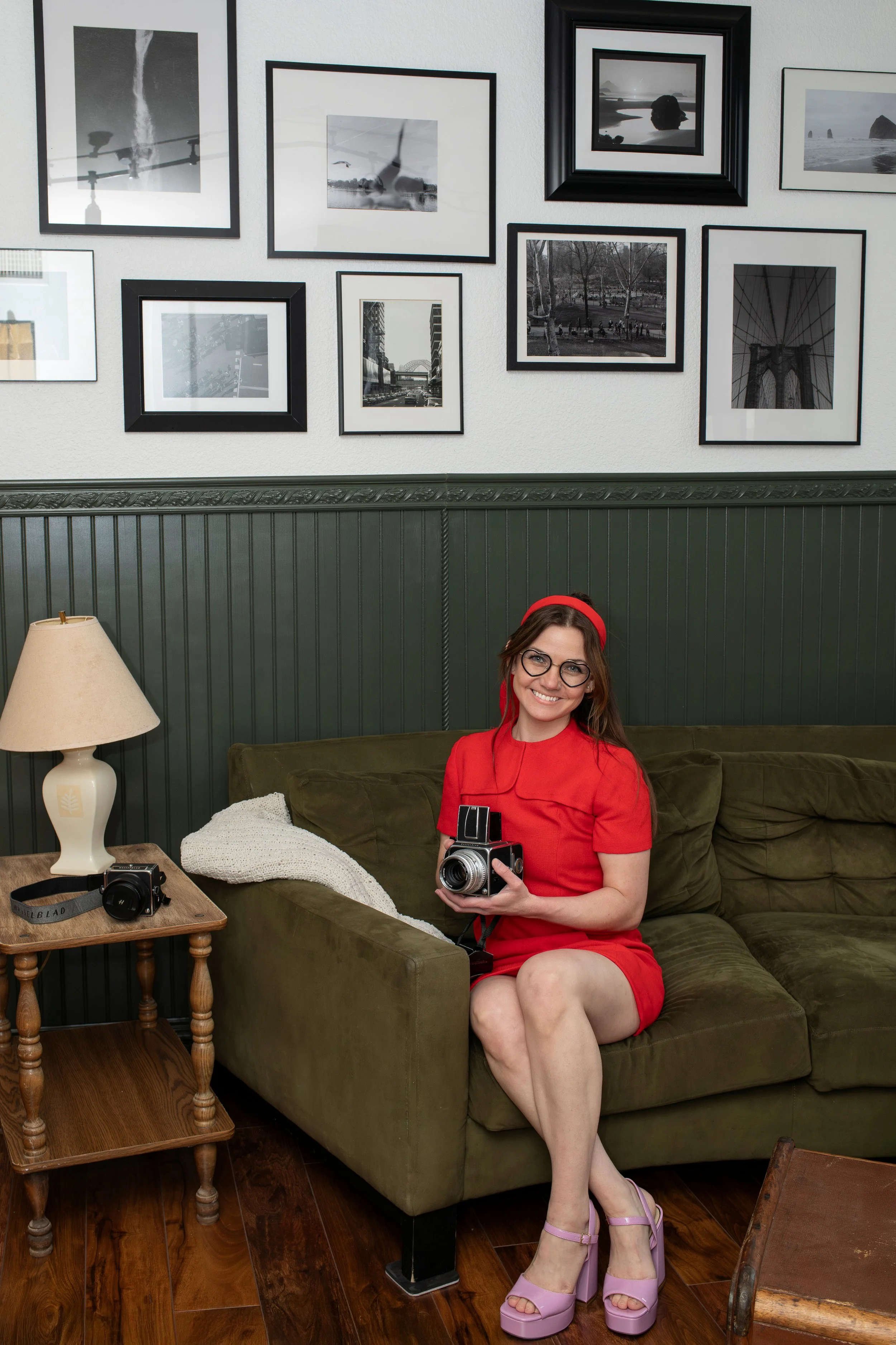 A woman with glasses, wearing a red dress and pink heels, sitting on a green sofa, holding a vintage camera, in a room with framed black-and-white photographs on the wall.